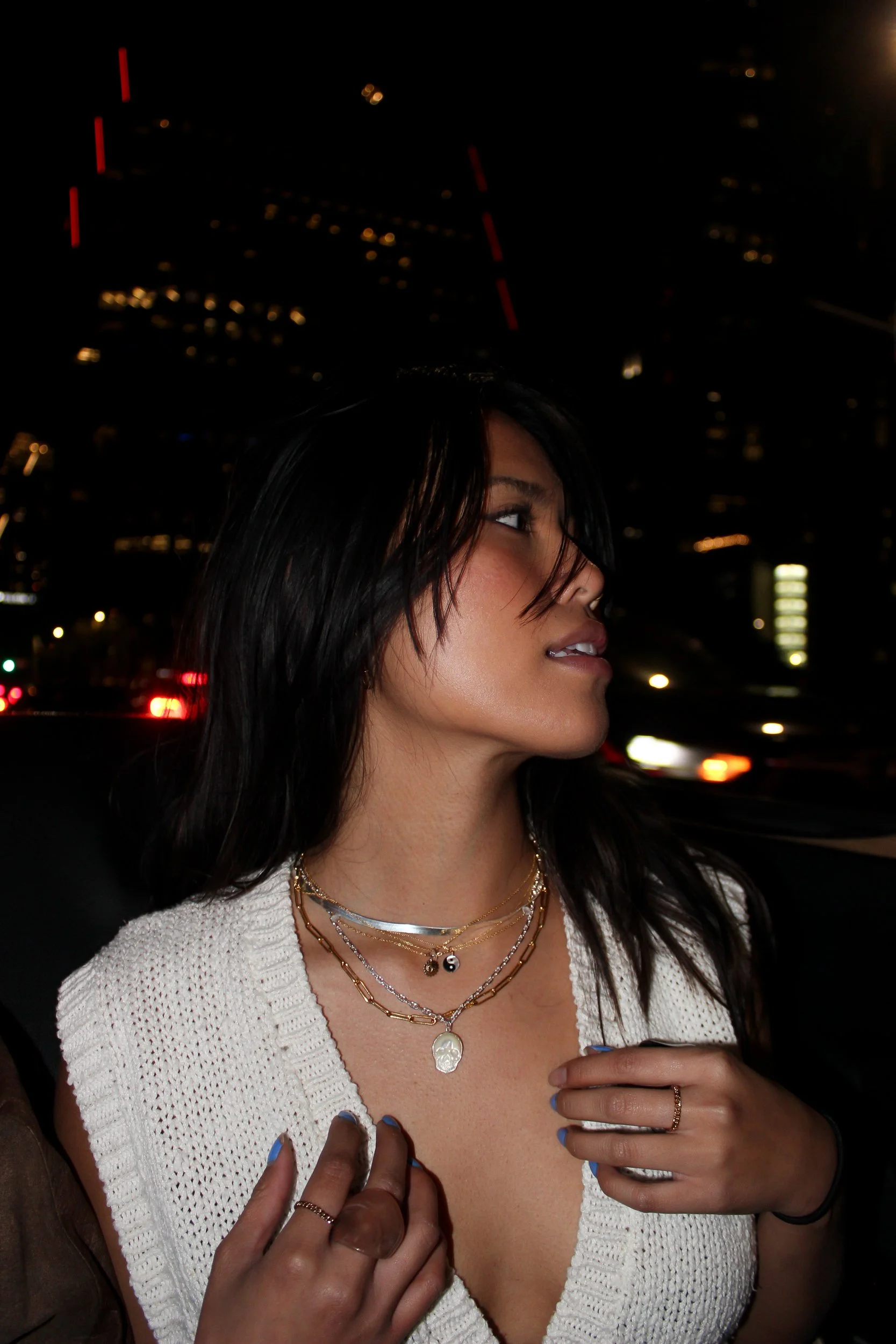 A woman with dark hair and layered jewelry, wearing a white knitted top, is shown sitting in a car at night. The city lights and buildings are visible in the background.