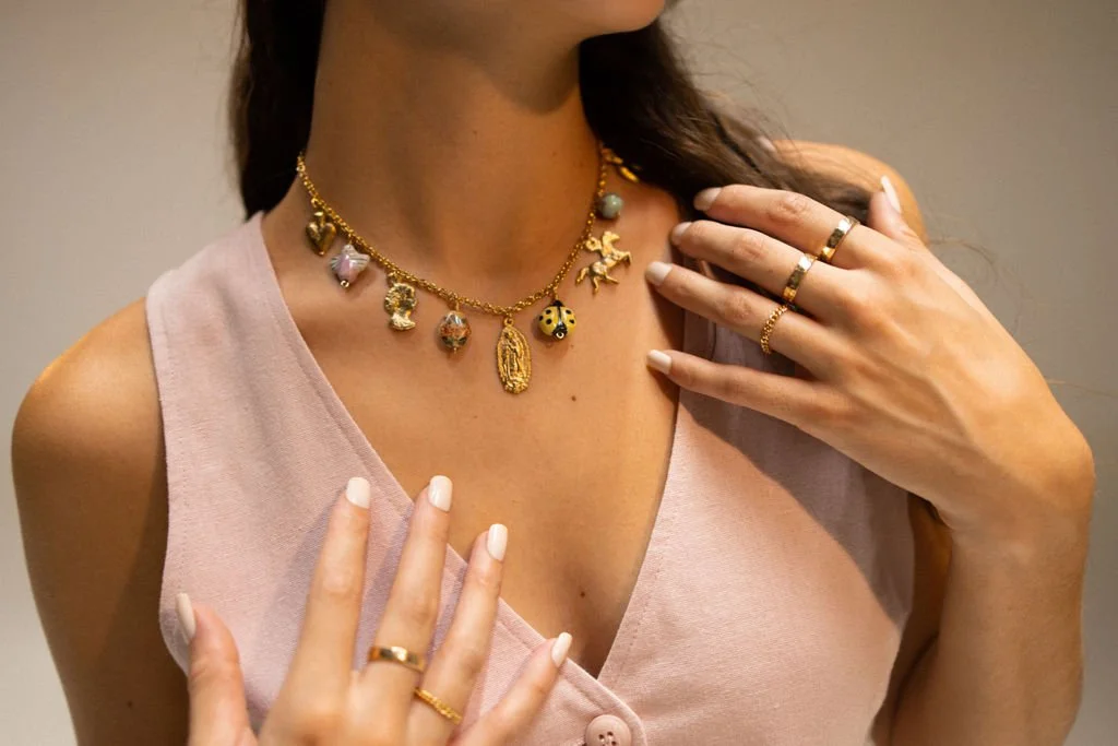 Close-up of a woman wearing layered gold necklaces with various charms, multiple rings on her fingers, and a sleeveless pink top.