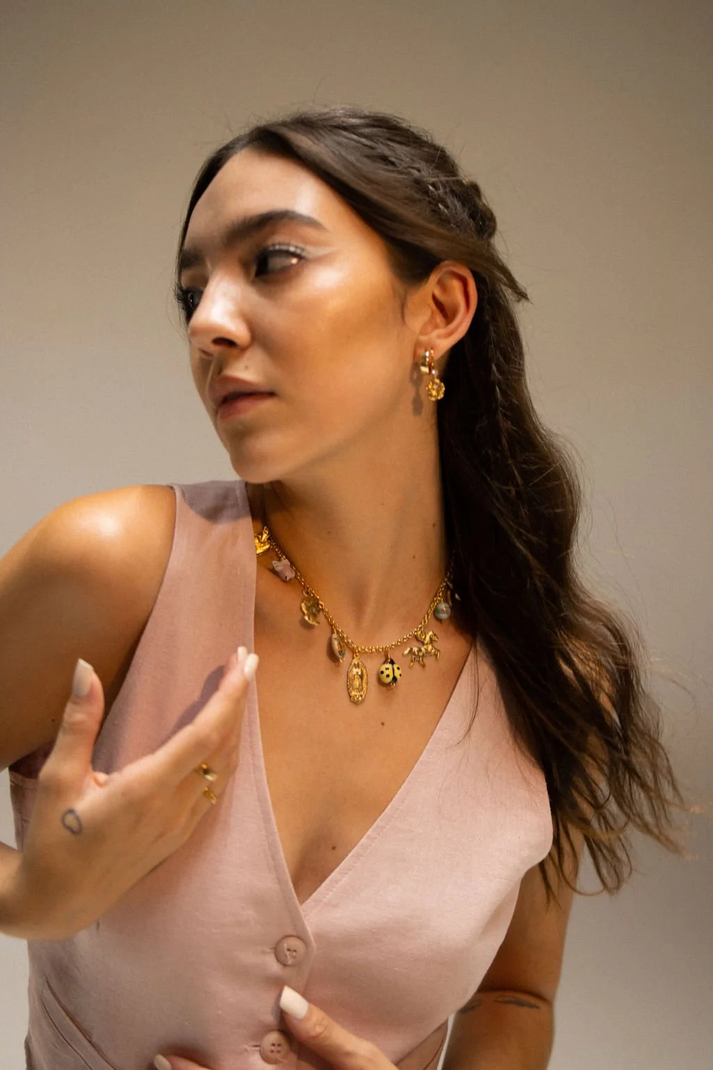 A woman wearing gold jewelry, including earrings and layered necklaces, with long wavy hair and wearing a pink sleeveless top.