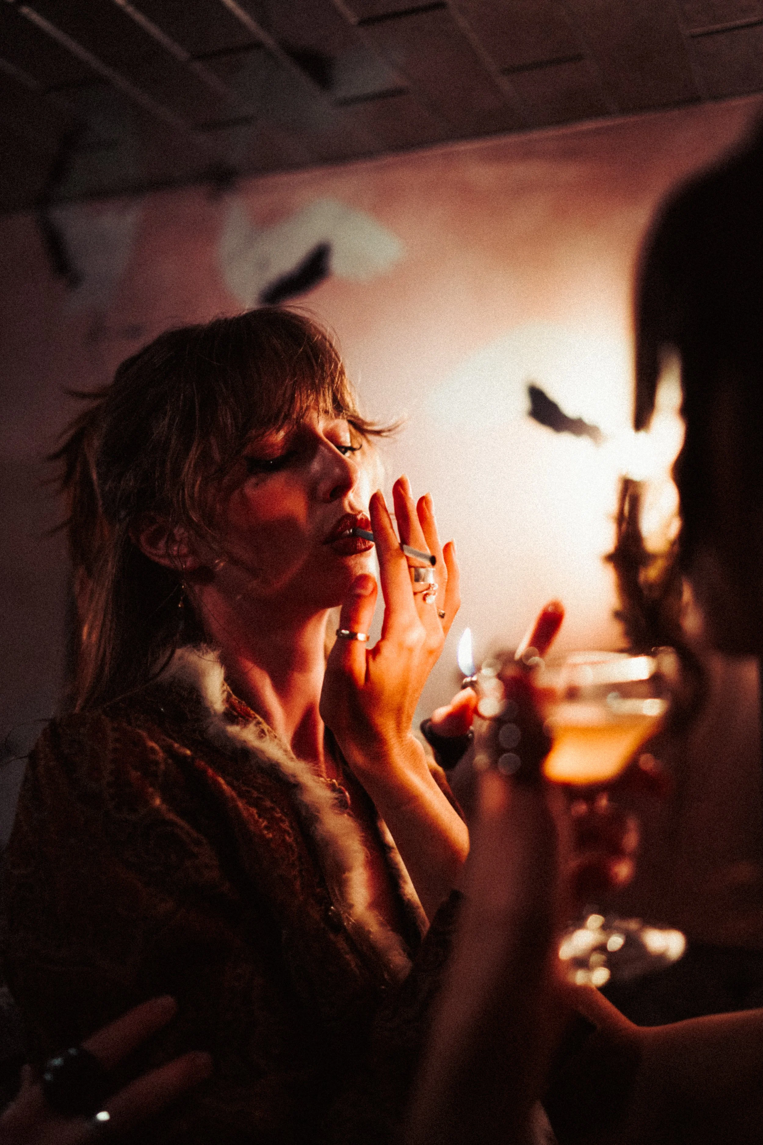 A woman with short, wavy hair smoking a cigarette in a dimly lit setting with warm, orange lighting. Her eyes are half-closed, and she is wearing rings on her fingers. There are candles and blurry figures in the background.