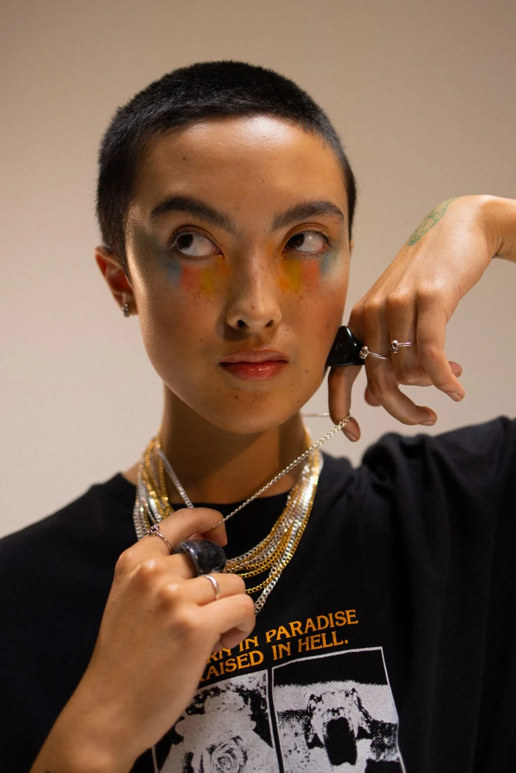 Person with short dark hair, colorful makeup, wearing layered jewelry, black T-shirt with a graphic and text, holding a chain necklace, against a plain background.