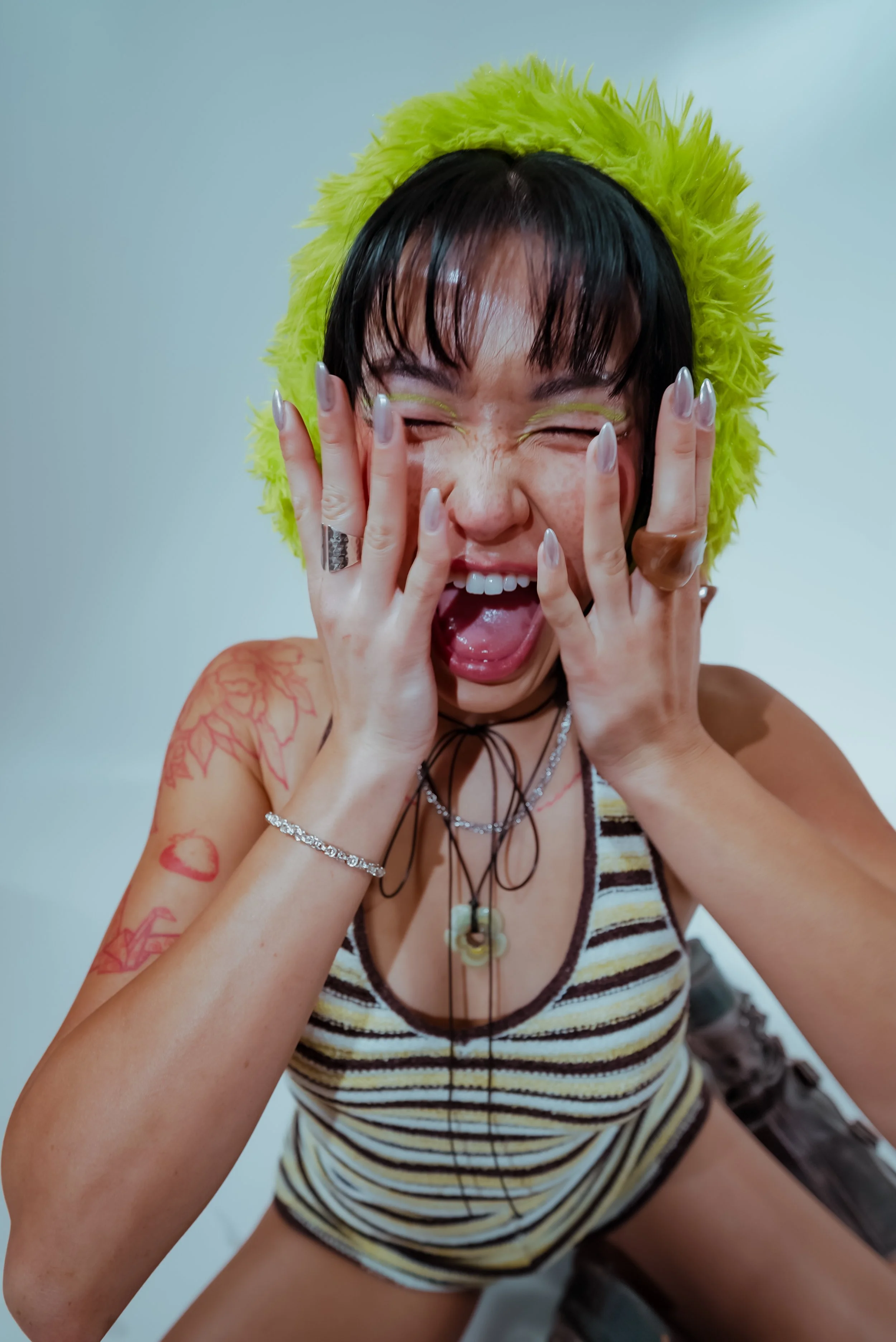 A woman with short dark hair and glasses, wearing a yellow furry hood, a striped tank top, and jewelry, laughing and covering her face with her hands, showing tattoos and colorful nail polish.