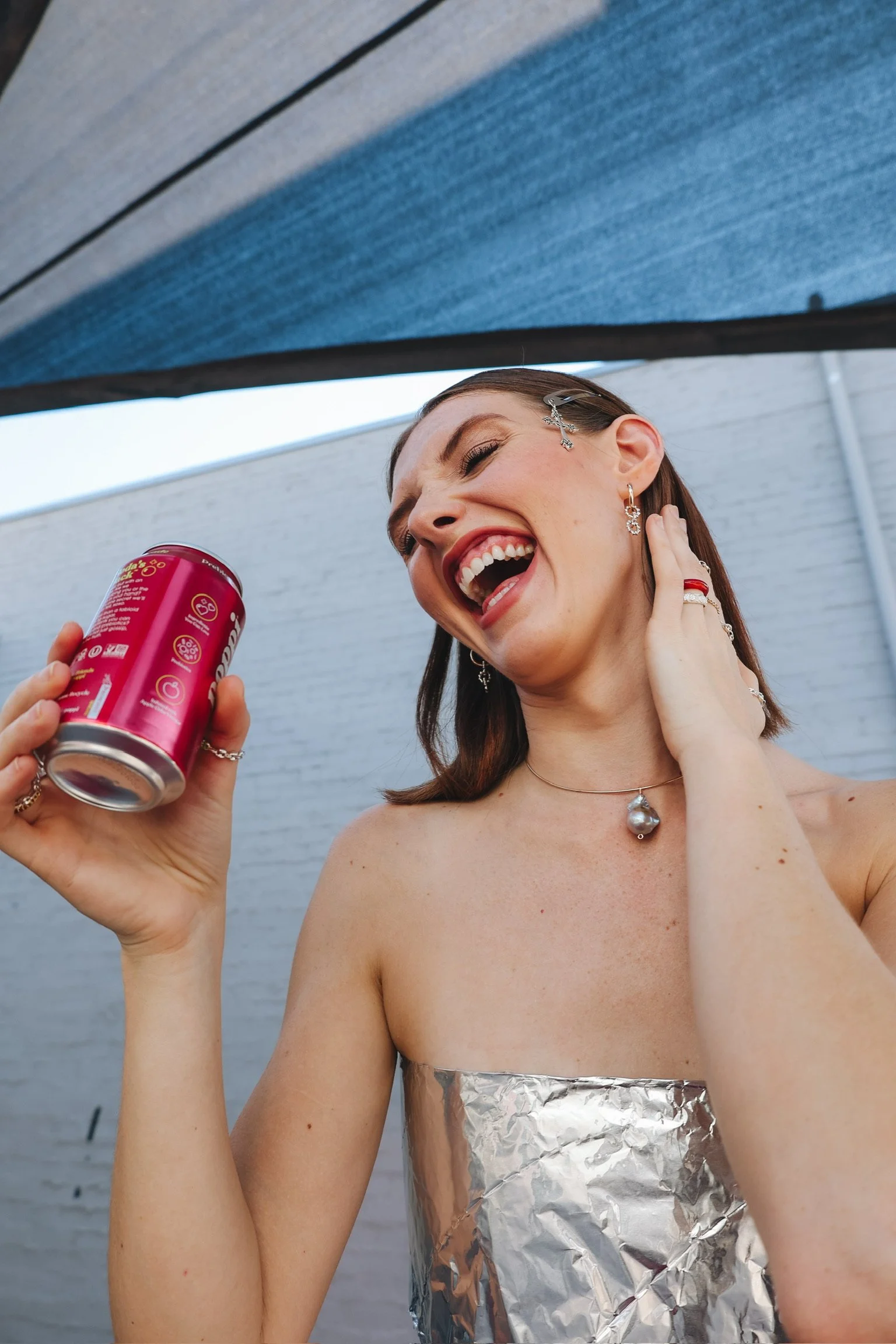 A woman with earrings and jewelry smiling, touching her ear, with a can of soda in a hand near her face, standing topless with aluminum foil covering her chest, outdoors under a shade with a brick background.