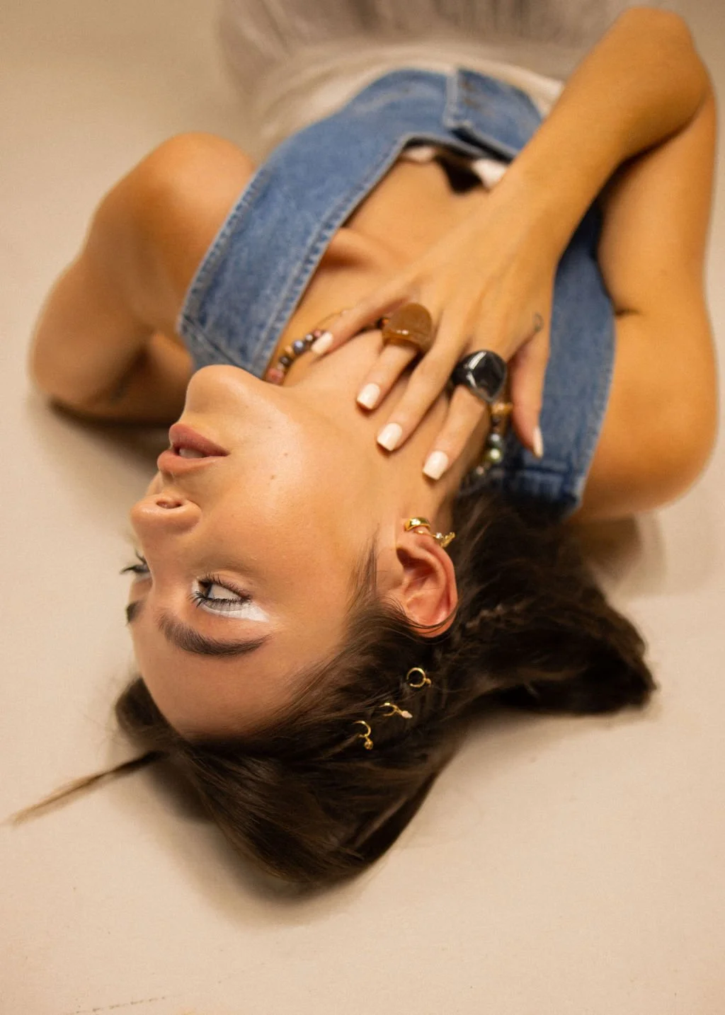 A woman lying on her side on a beige floor, wearing a denim vest and jewelry, with her hand touching her face and her eyes closed.