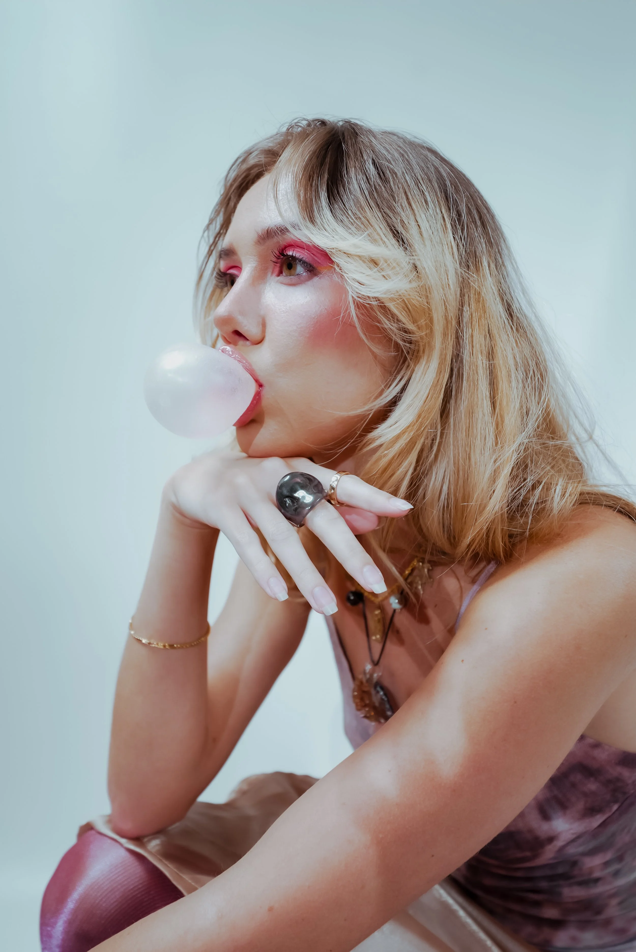 A woman with blonde hair and pink makeup blowing a bubblegum bubble, sitting with her chin resting on her hand, wearing a purple dress and accessories.