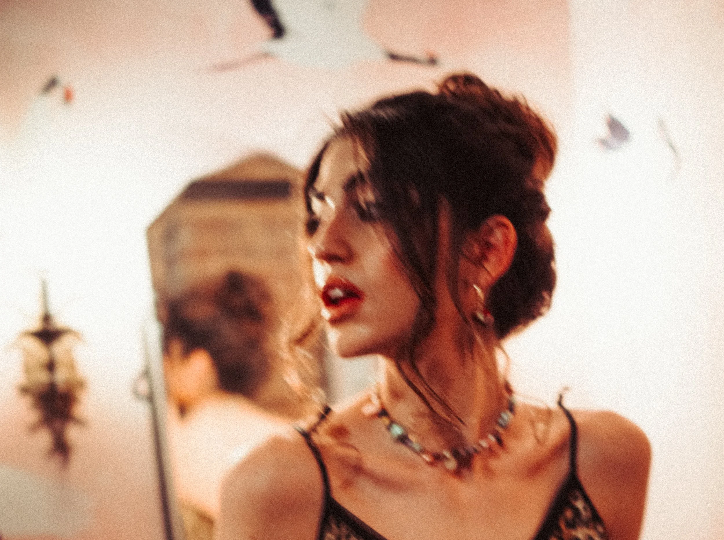 A young woman with dark hair, glasses, and red lipstick, wearing a black bra and jewelry, standing outdoors at sunset with a blurred background of streetlights and sky.