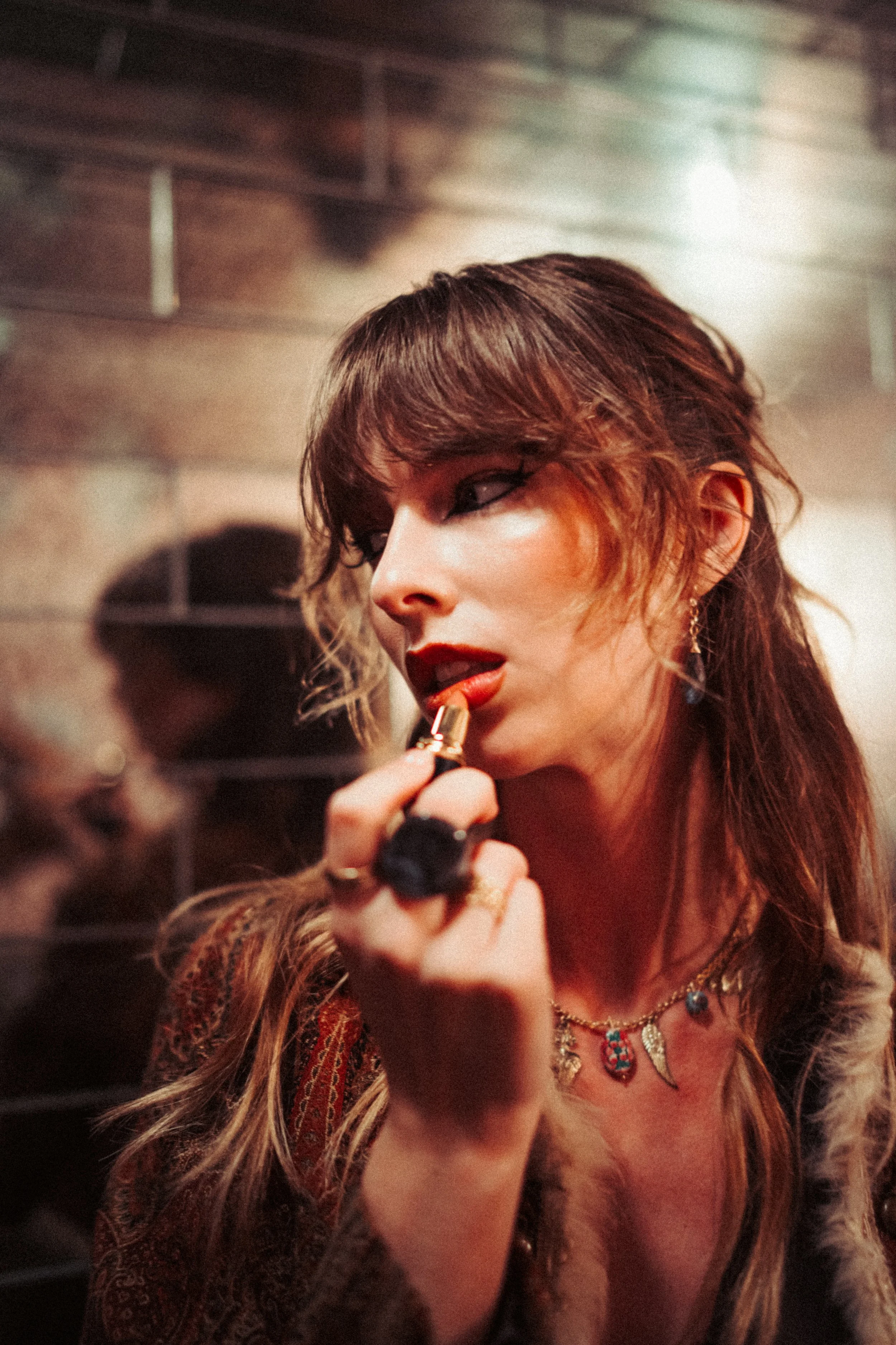 A woman with light skin and long, wavy hair with bangs is applying red lipstick while looking into a mirror. She is wearing a leopard print top, layered necklaces, and earrings. The background appears to be brick or tiled wall with soft lighting.