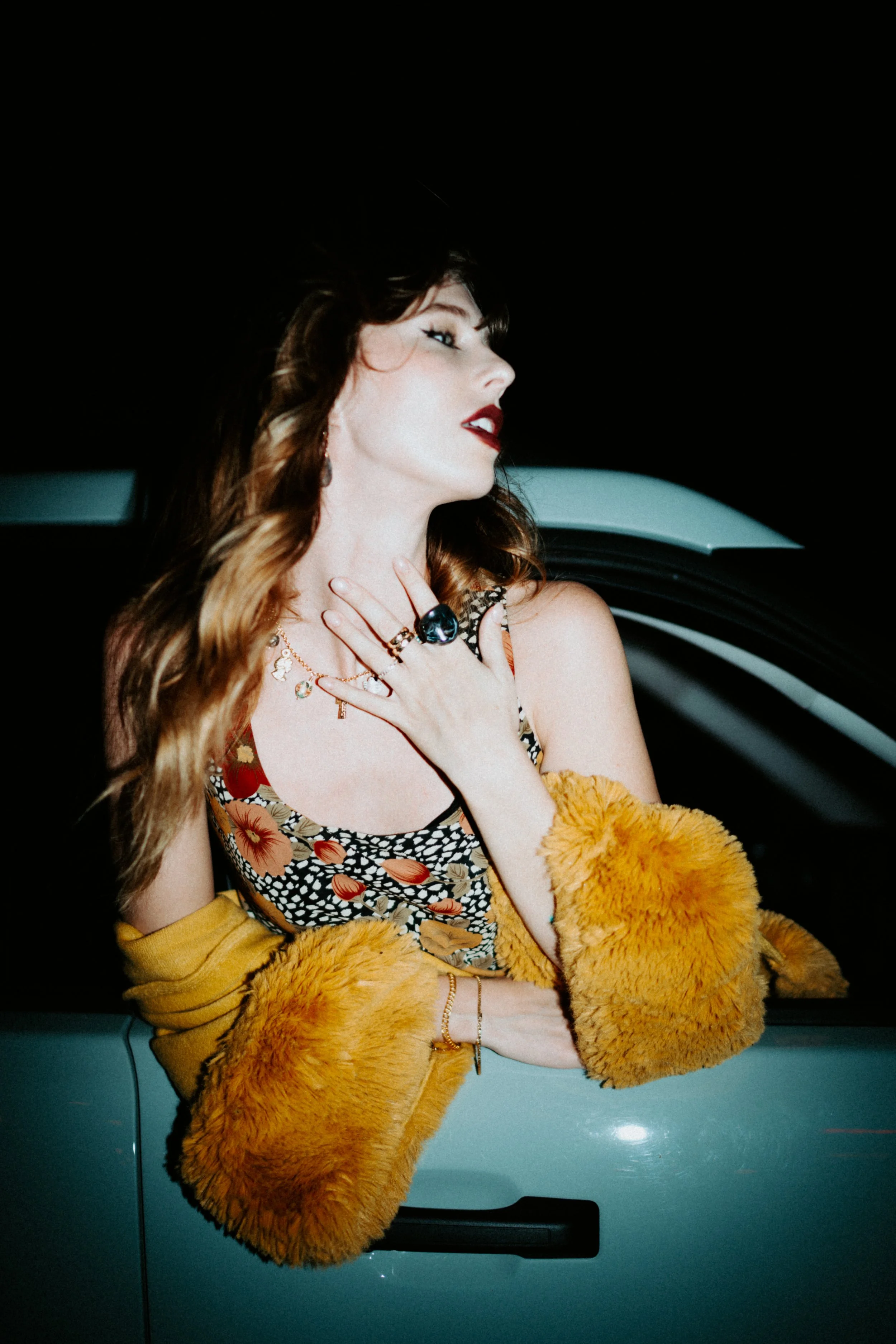 A woman with brown and blonde hair wearing red lipstick, gold jewelry, a floral dress, a yellow fur coat, posing with her head tilted back and hand on her neck, standing in front of a dark background and a blue vehicle.