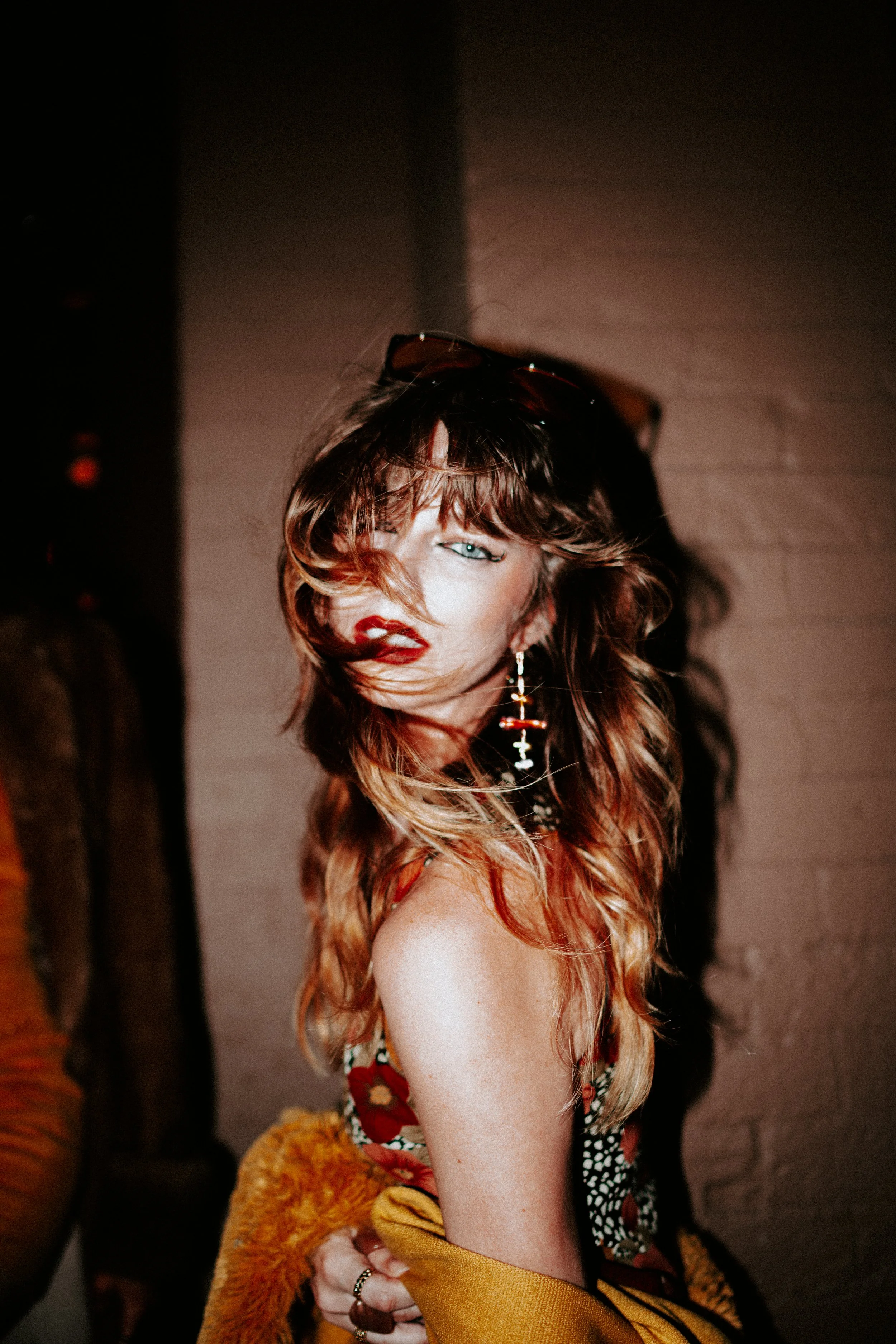 A woman with long wavy hair, partially covering her face, wearing sunglasses on her head, a black and white patterned dress, and has red lipstick. She is holding a fluffy yellow-orange coat and has a cross earring in her left ear.