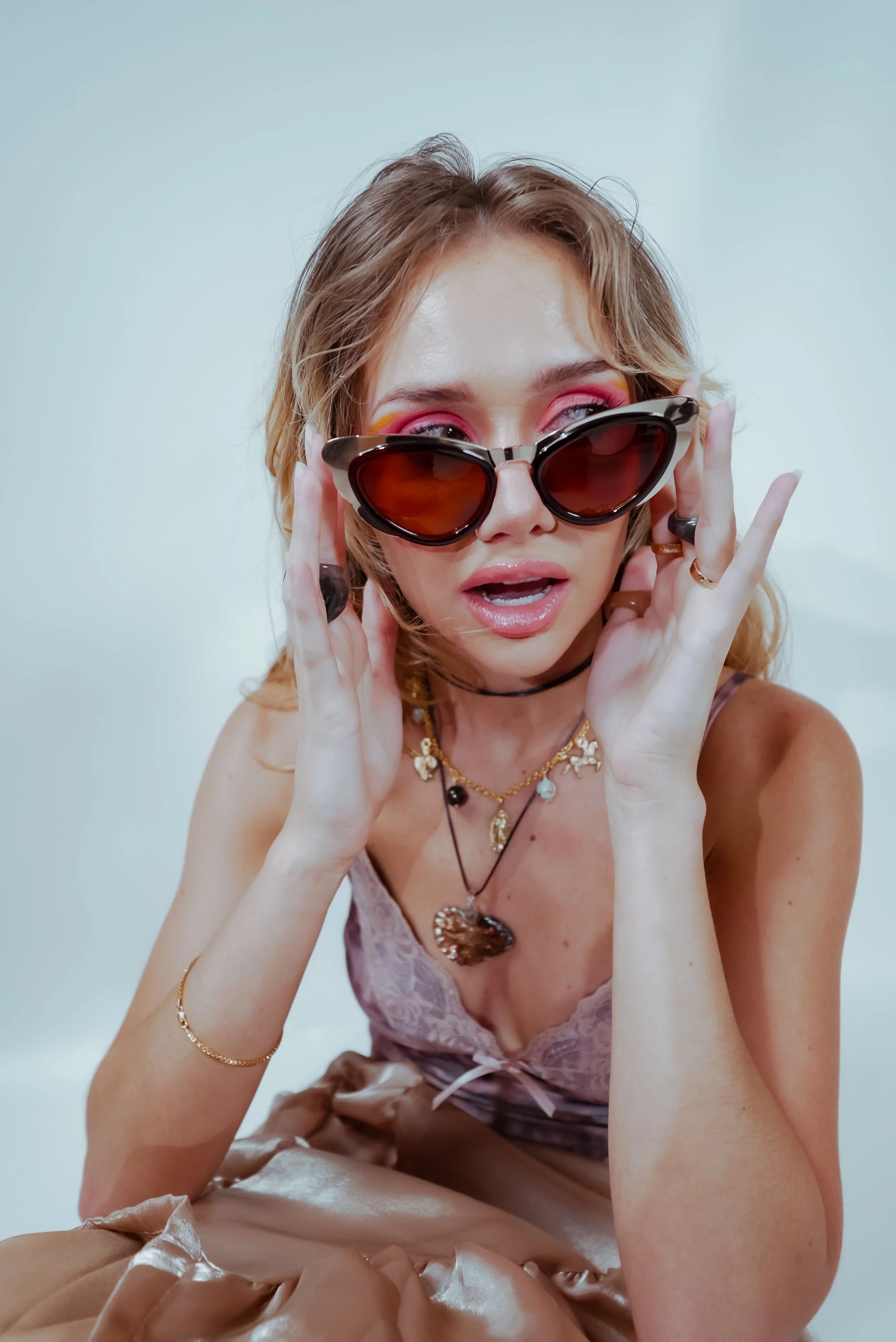 A woman with light brown, shoulder-length hair wearing large cat-eye sunglasses, pink eye makeup, and jewelry including necklaces and rings, sitting against a plain white background.