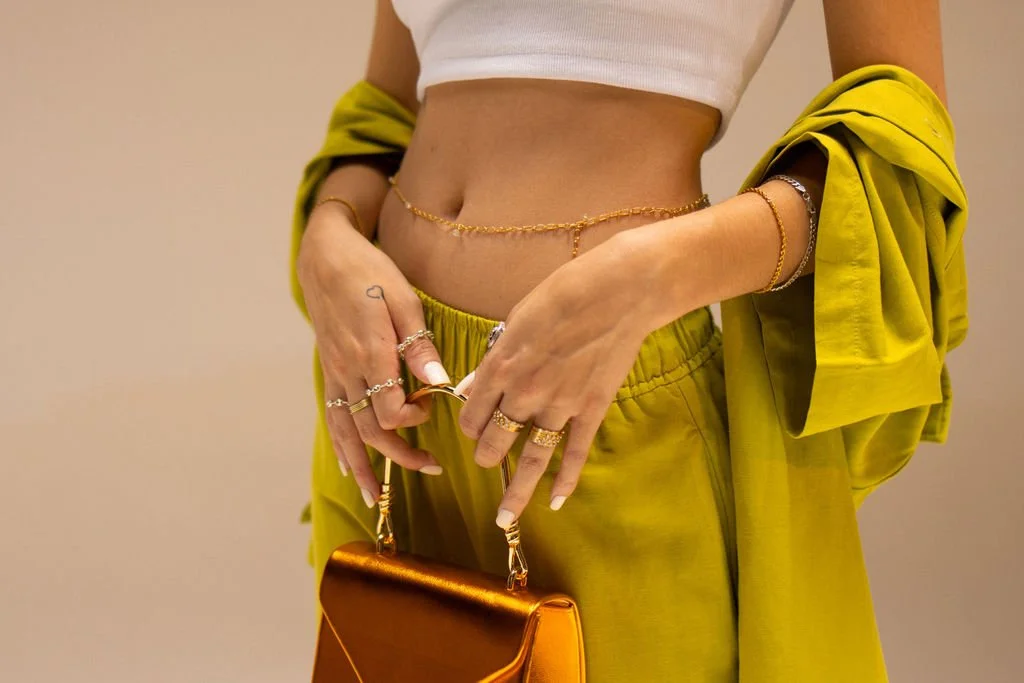 A woman in a white crop top and matching lime green pants holding a metallic orange clutch, with her hands adorned in rings and bracelets, and a yellow jacket draped over her arm.