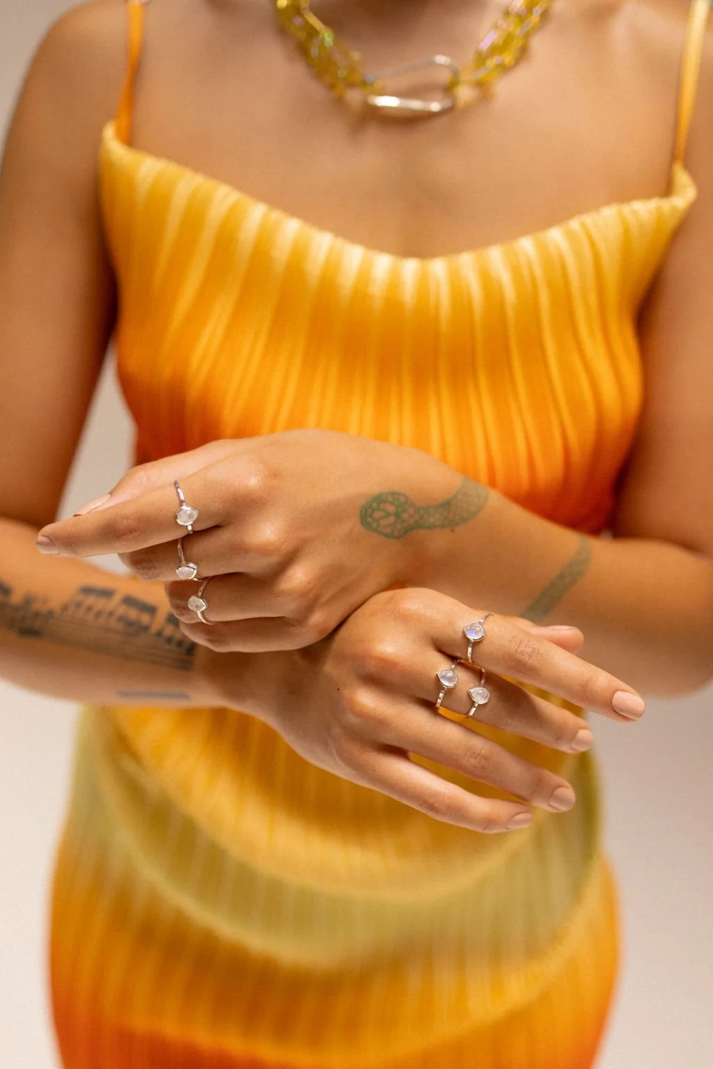 A woman wearing a yellow and orange striped dress, gold jewelry, and multiple rings, with tattoos visible on her arms and hands.