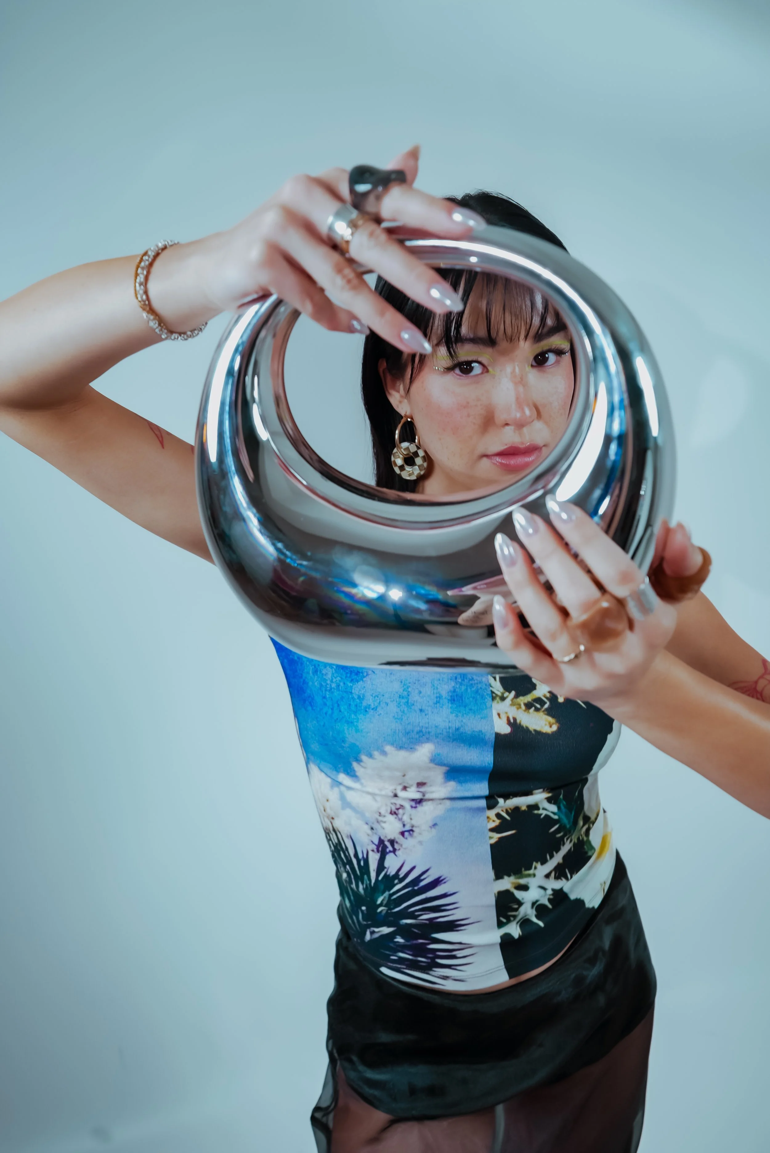 A woman with black hair and earrings holding a mirror, looking into it. She is wearing a colorful, artistic top and a sheer black skirt.