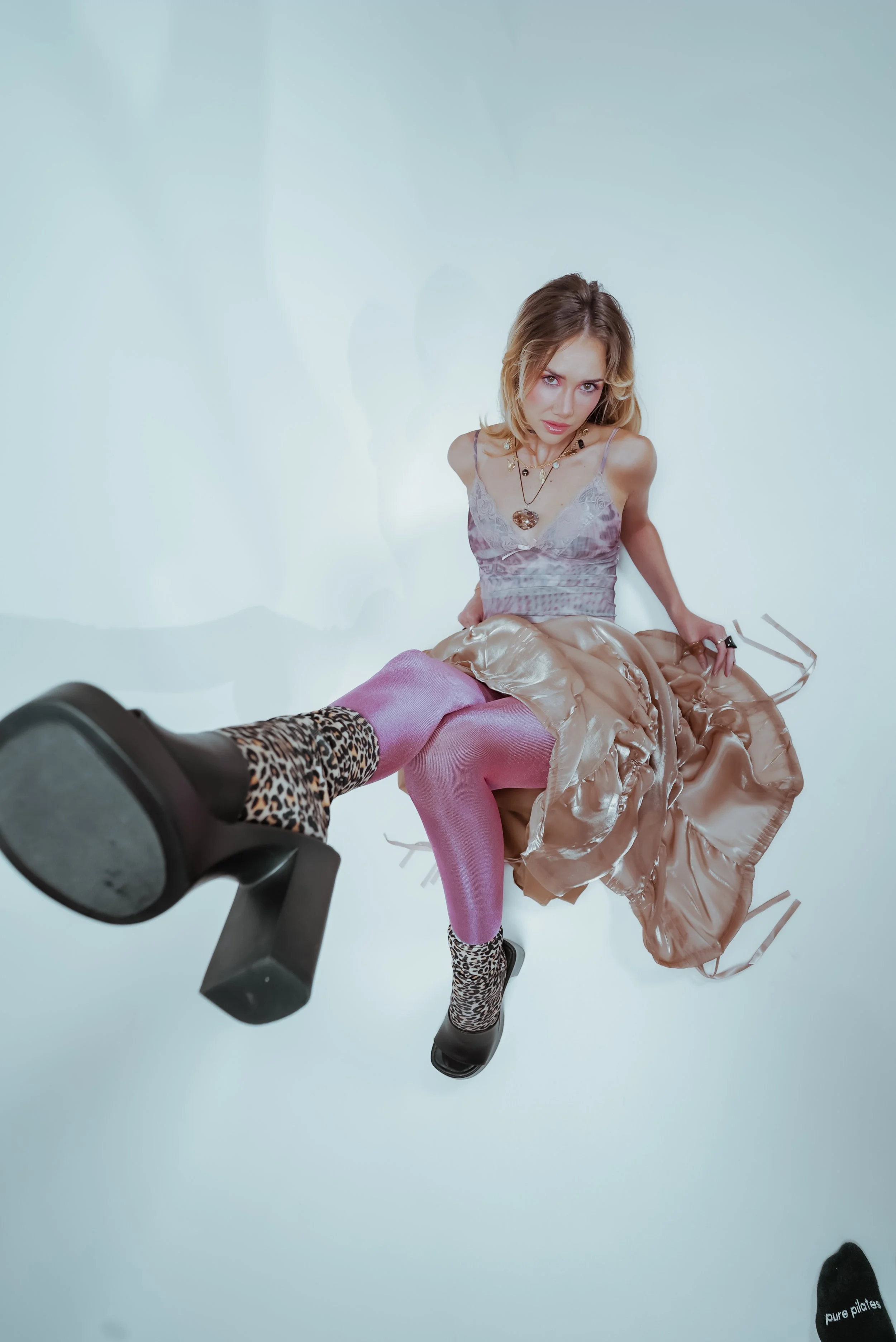 A woman with blond hair wearing a purple top, pink tights, leopard print socks, and leopard print shoes, sitting on a silver-colored, shiny dress, with her legs extended and looking at the camera against a plain white background.