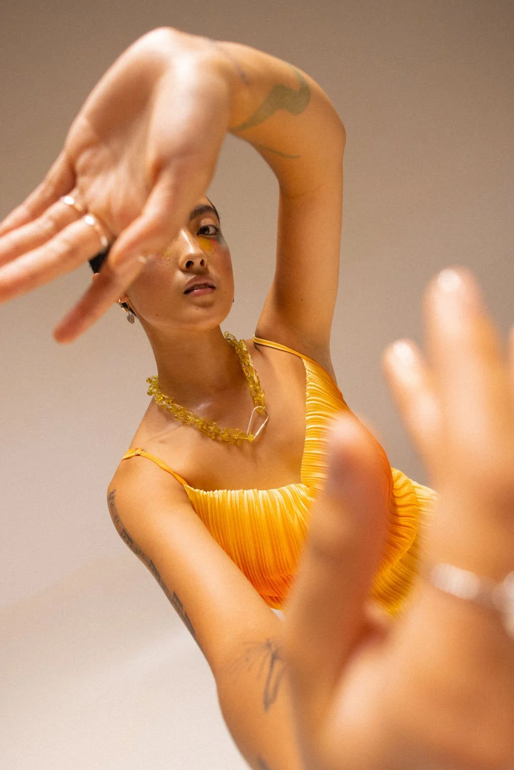 A woman in a yellow dress making a hand gesture, with an expression looking at the camera, standing against a plain background.