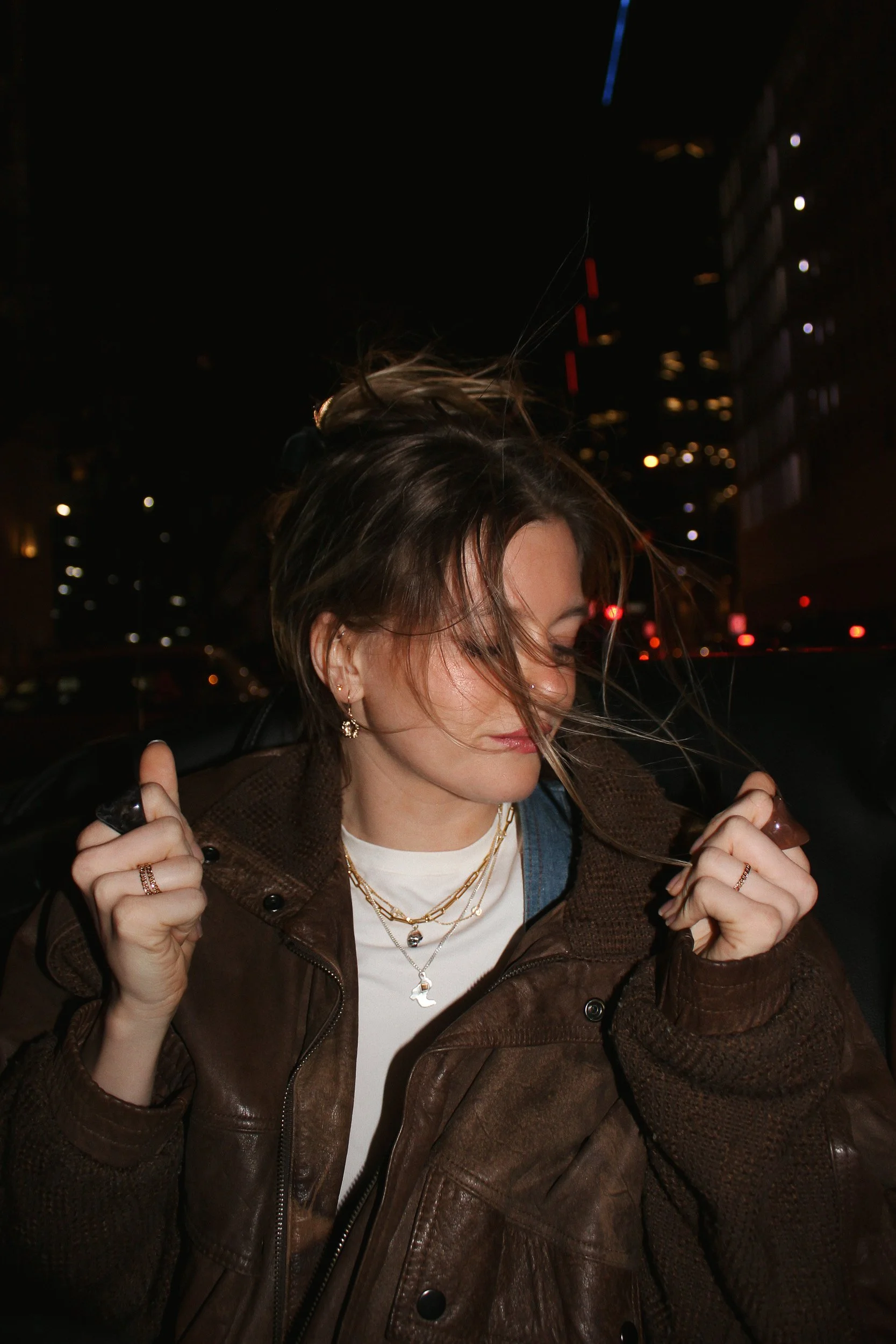 A woman with shoulder-length hair, wearing a brown leather jacket, multiple silver necklaces, and earrings, stands outdoors at night with city buildings in the background. Her hair is being blown by the wind and she appears to be holding sunglasses i