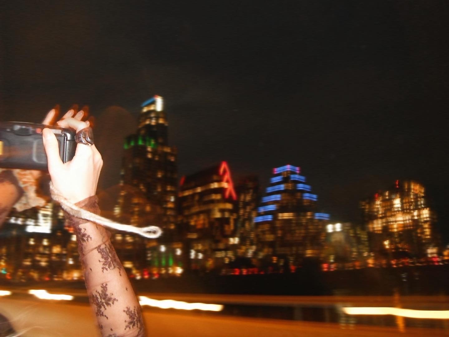 Person with a lace sleeve taking a photo of a city skyline at night, with illuminated buildings and blurred lights in the background.