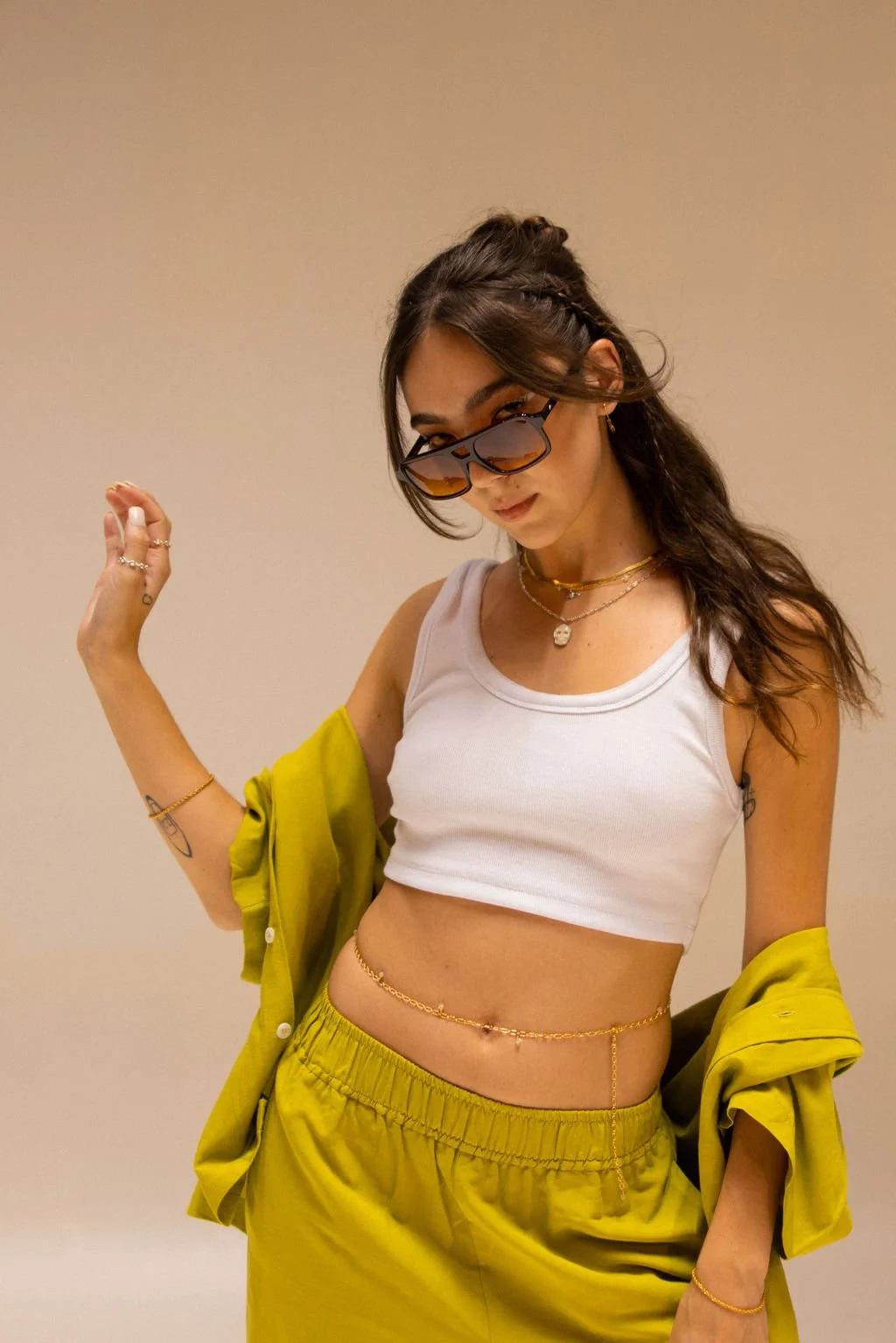 Young woman wearing sunglasses, white crop top, yellow pants, and a yellow shirt draped over her shoulders, accessorized with jewelry.