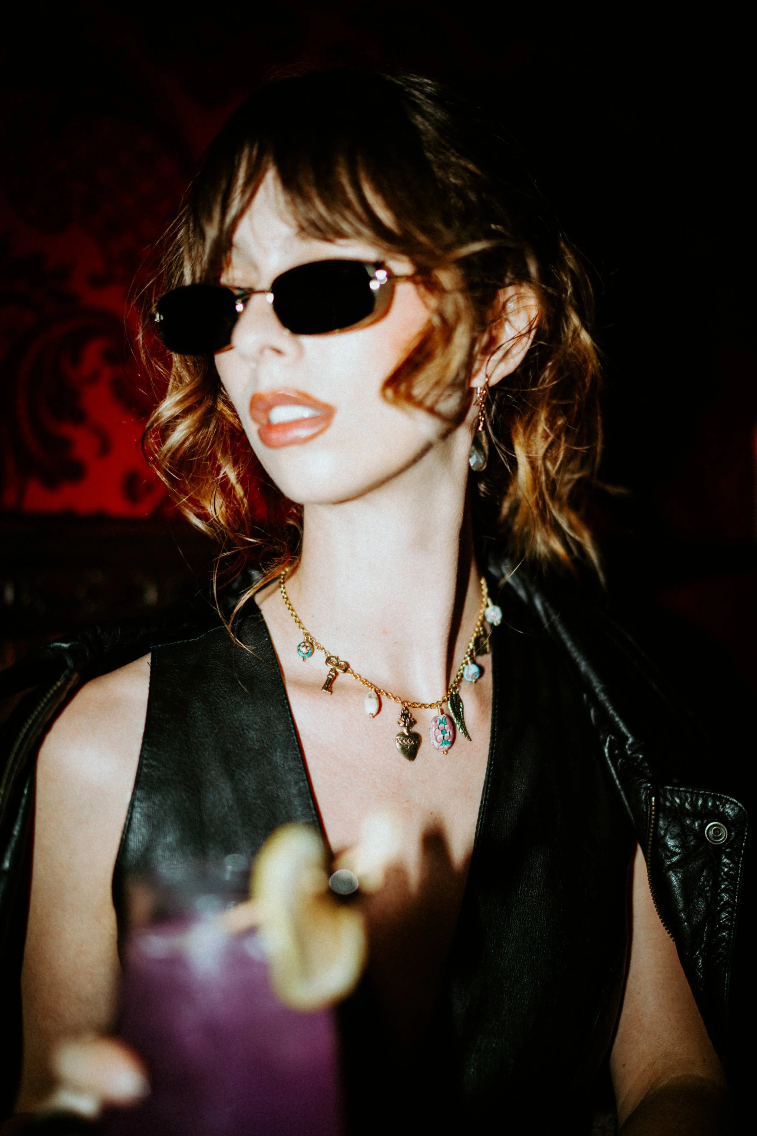A woman with wavy brunette hair and sunglasses, wearing layered necklaces and a sleeveless black leather jacket, appears to be at a social event or party with red and black decor in the background.
