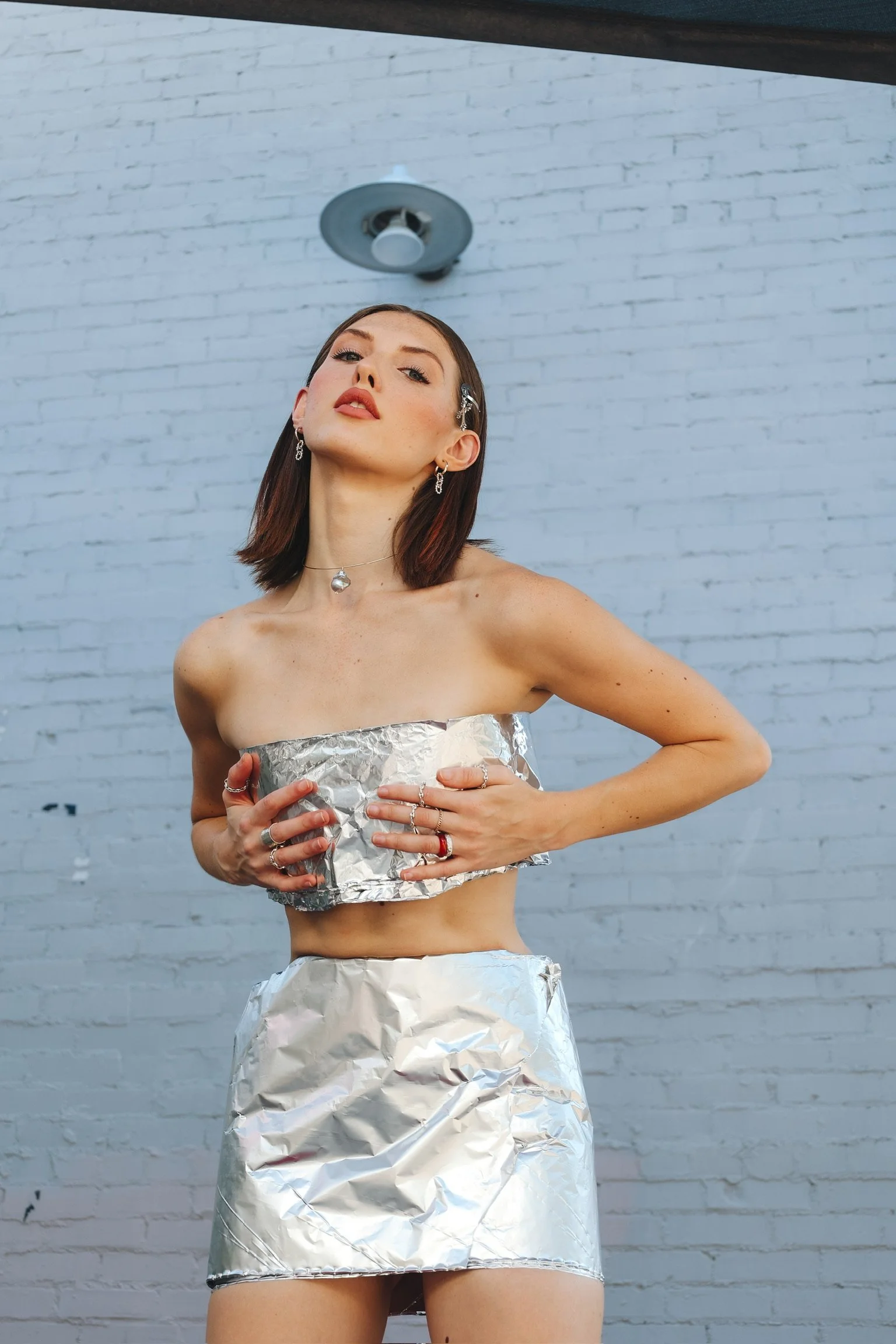 A young woman with shoulder-length brown hair and multiple earrings poses with her head tilted slightly upward in front of a white brick wall. She is wearing a strapless top and skirt made of crumpled aluminum foil, holding the foil fabric in her hands.