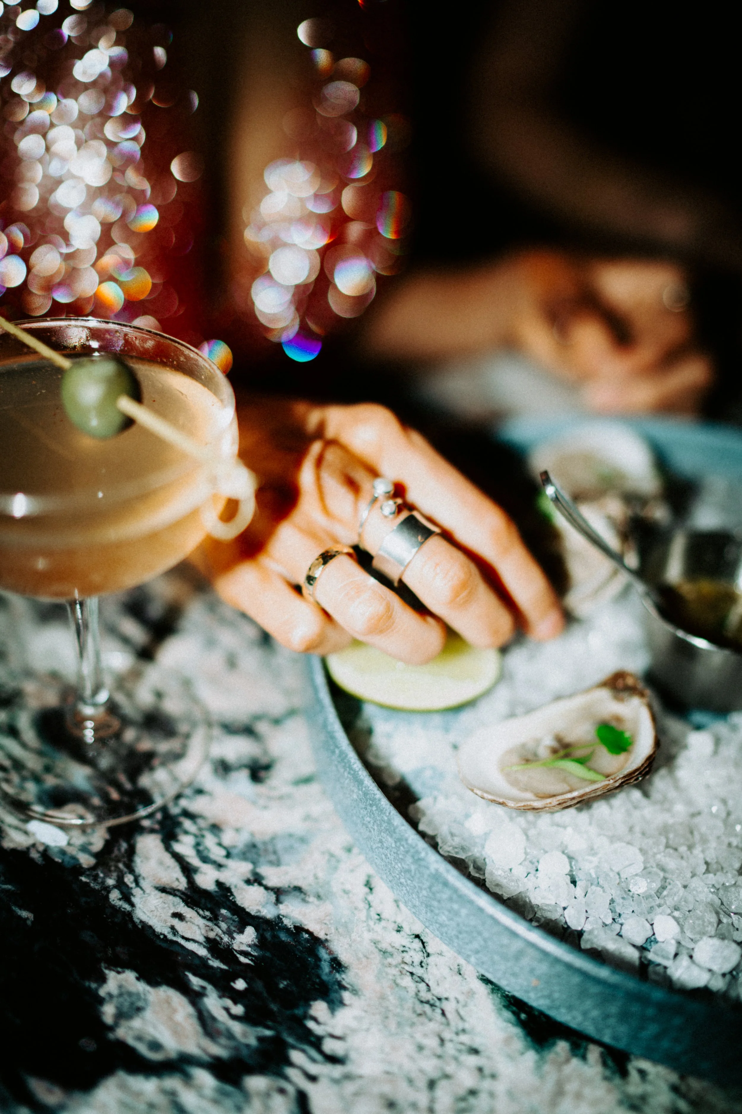 Close-up of a seafood platter featuring oysters on the half shell with garnishes, a cocktail glass with a green olive, and a person's hand with multiple rings, all on a marbled countertop with colorful bokeh lights in the background.