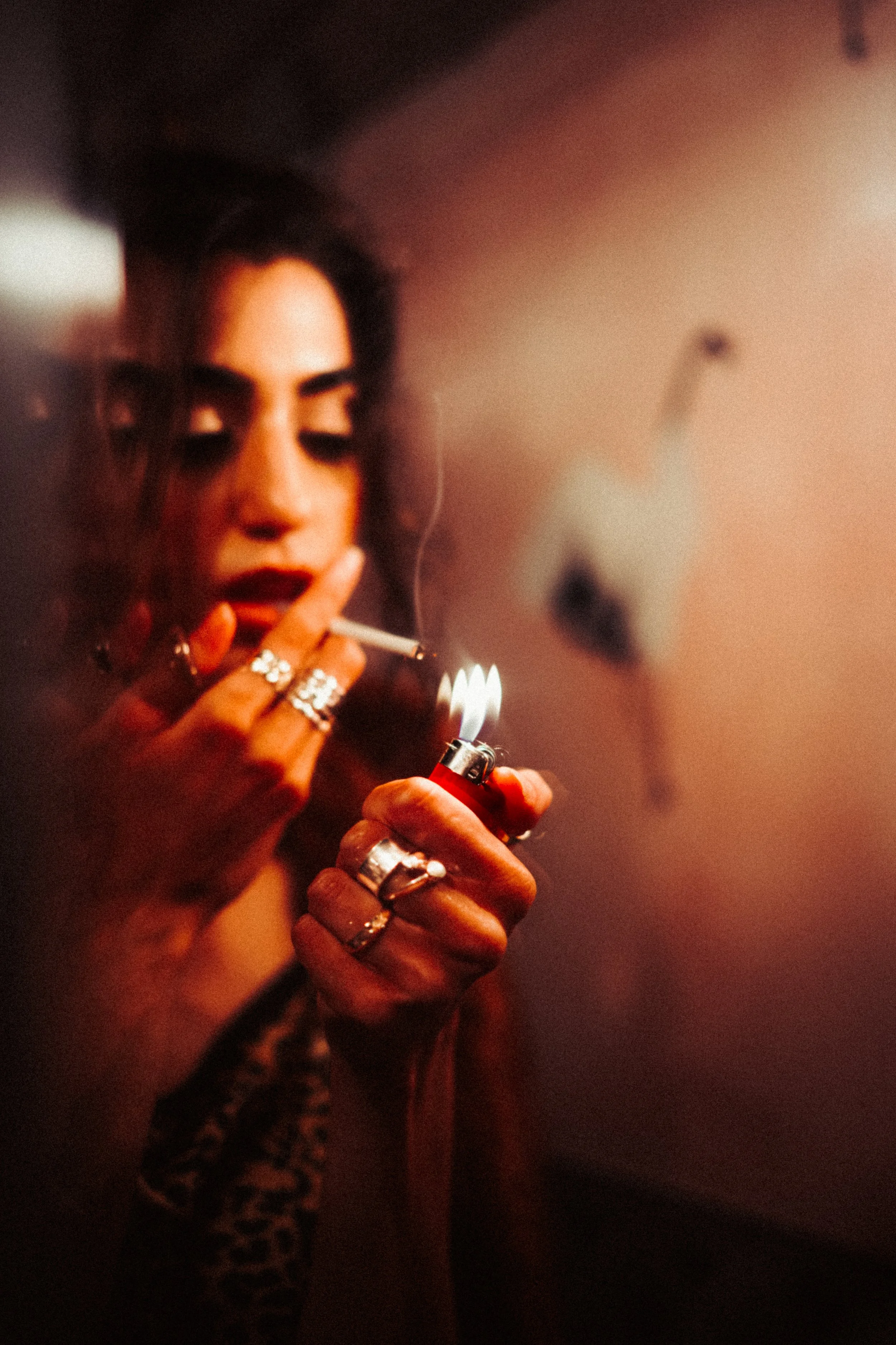A person with dark hair and multiple rings on fingers is lighting a cigarette with a red lighter in a dimly lit environment.