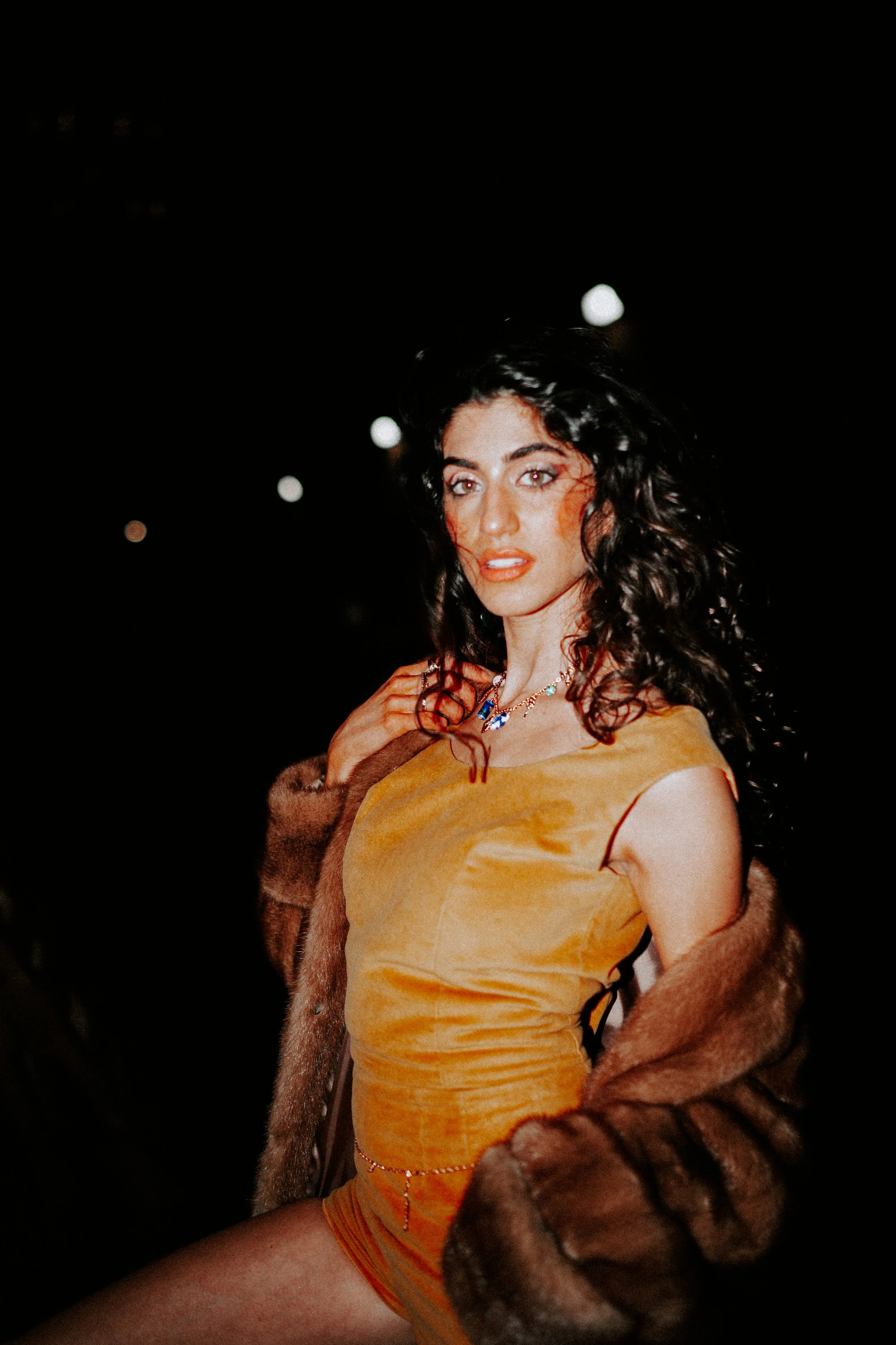 A woman with dark curly hair wearing a yellow dress, a pearl necklace, and a fur coat, standing against a dark background with blurred lights.