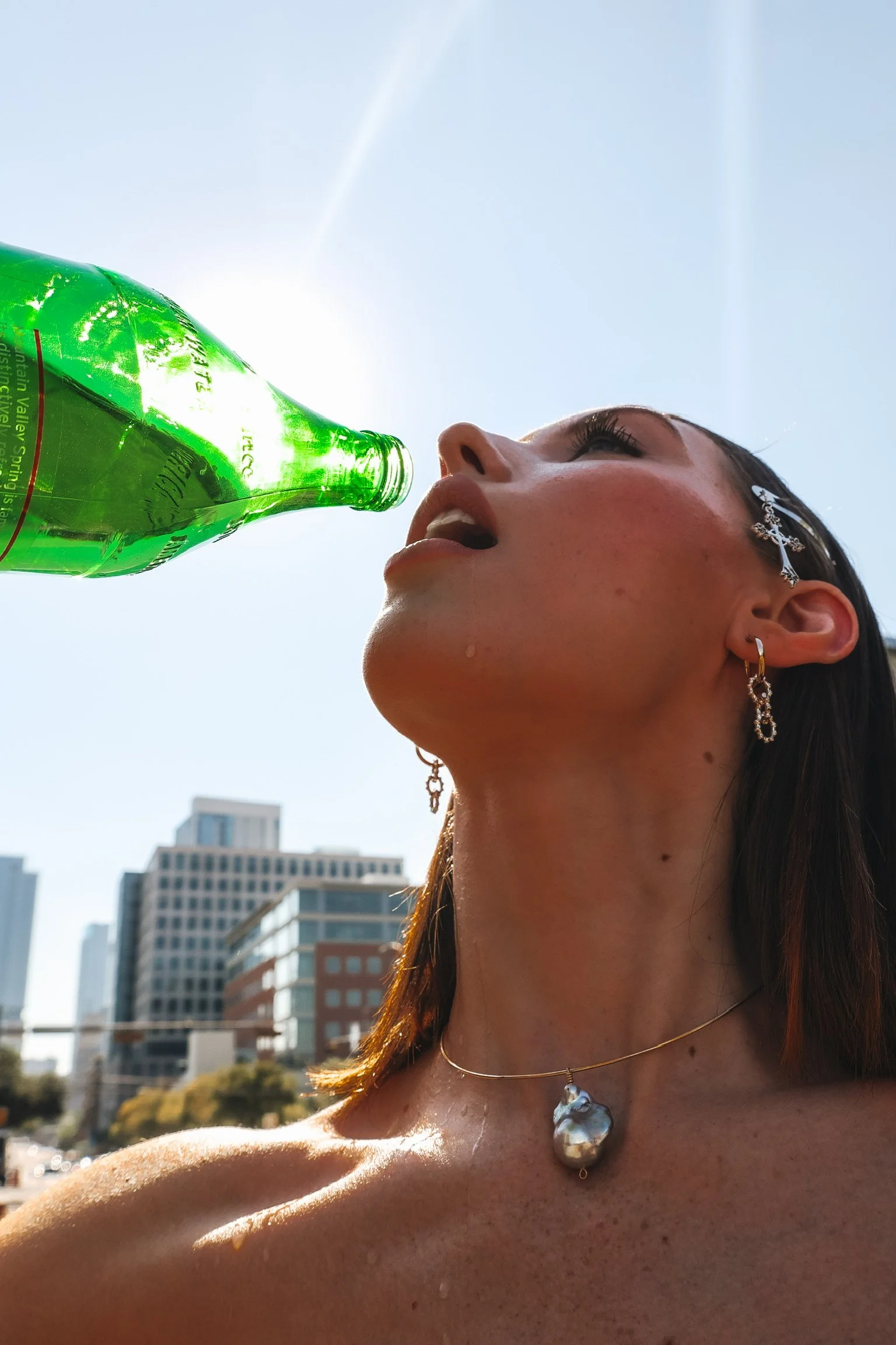 A woman with earrings and a necklace, standing outdoors on a sunny day in a city, drinking from a green glass bottle.