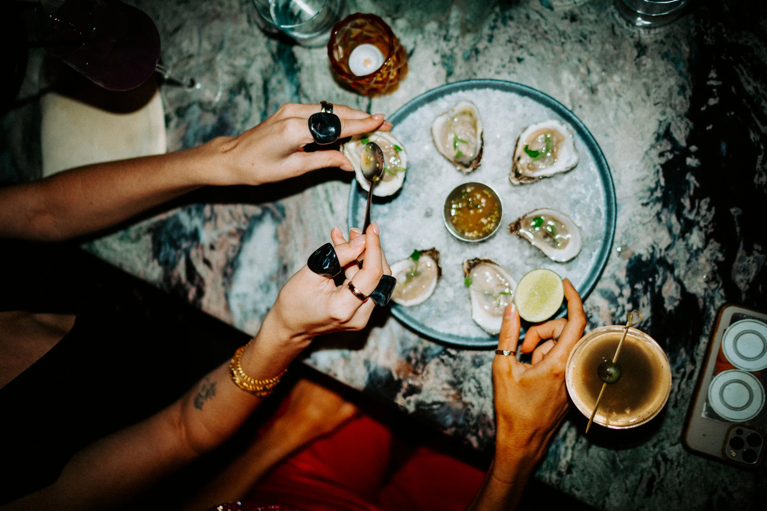 People sharing oysters, with one person squeezing lemon over them and another holding a drink with a lime, on a marble table with drinks and food.