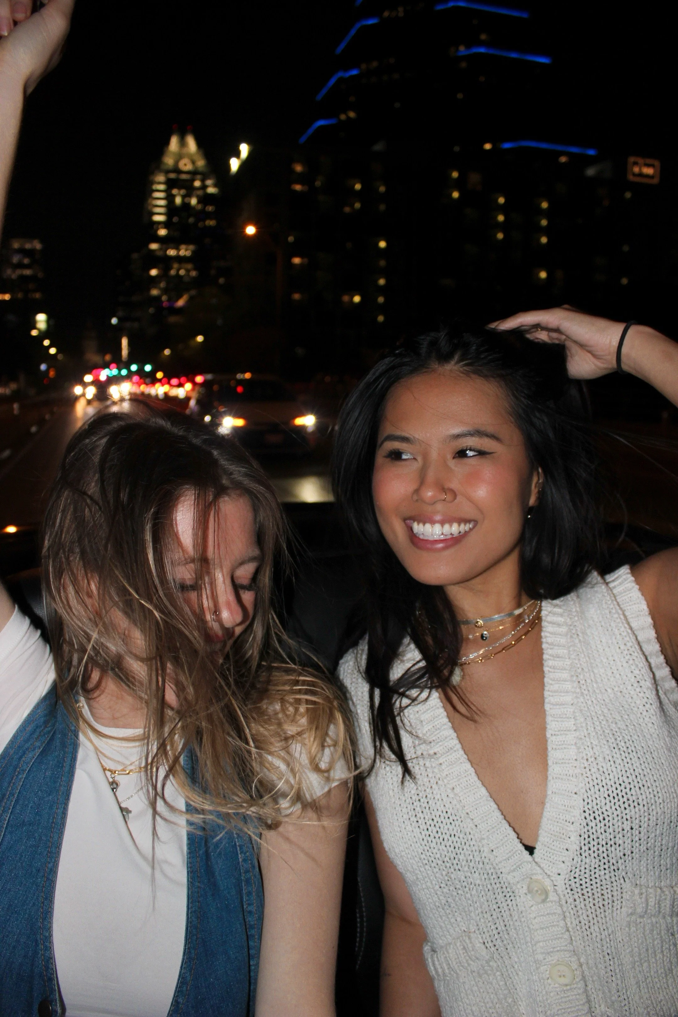 Two women smiling at night with city buildings and lights in the background. One woman has long, wavy, blonde hair and is wearing a white shirt with a denim vest. The other woman has straight, black hair and is wearing a white, knitted top.