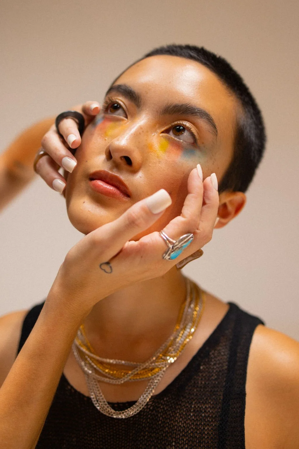 A person with short dark hair and gold jewelry has colorful makeup around their eyes, and a hand with rings gently touches their face.