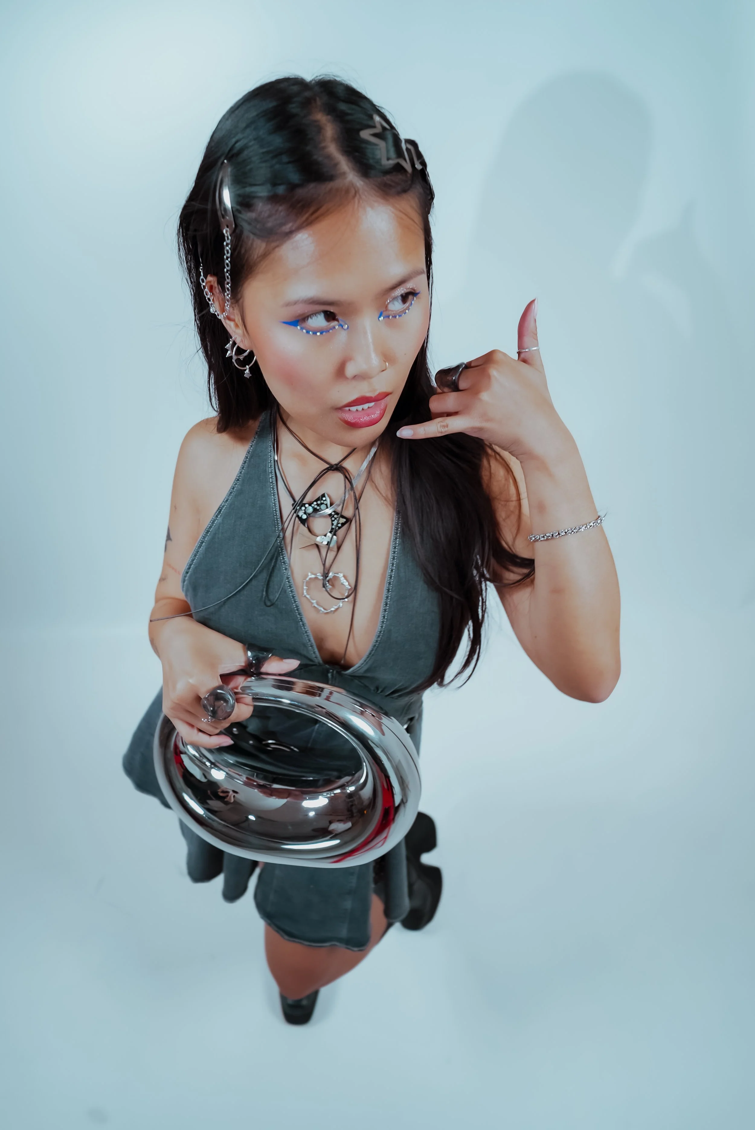 A young woman wearing a gray dress, with dark hair styled with star-shaped hair clips, holding a metallic mirror and making a phone gesture with her right hand. She has several accessories, including layered necklaces, earrings, and rings, and has bl