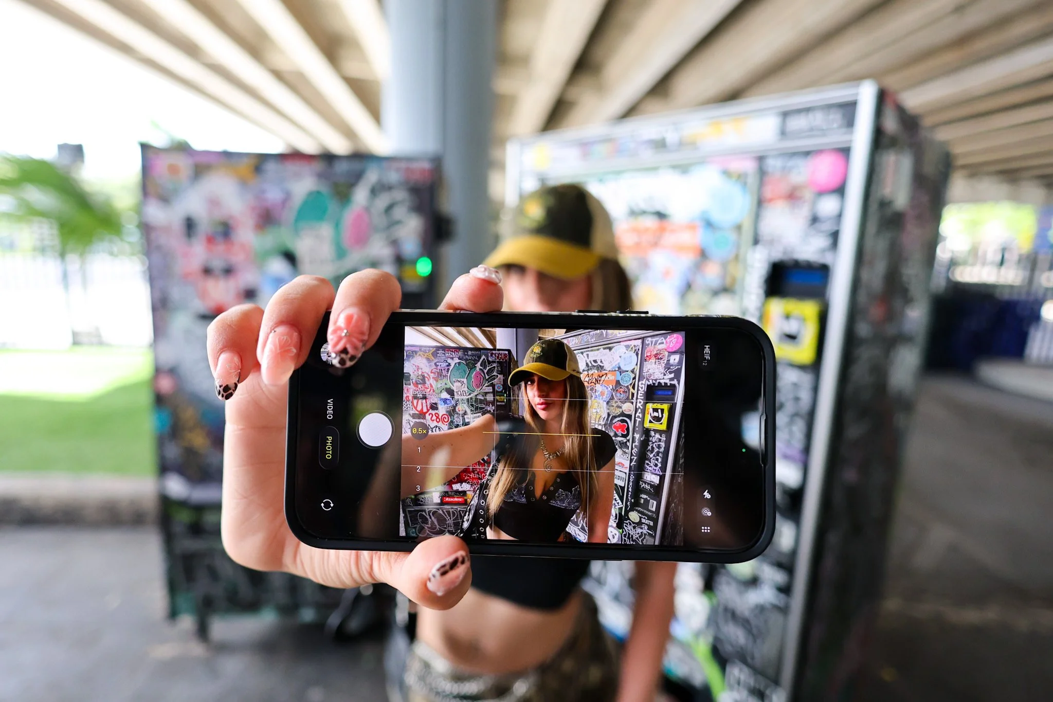 A person with long hair and a yellow cap is taking a selfie with a smartphone in front of a graffiti-covered wall under a bridge or overpass. The photo shows the person on the phone's screen with the graffiti in the background, while the person’s face is slightly blurred outside the phone's screen.