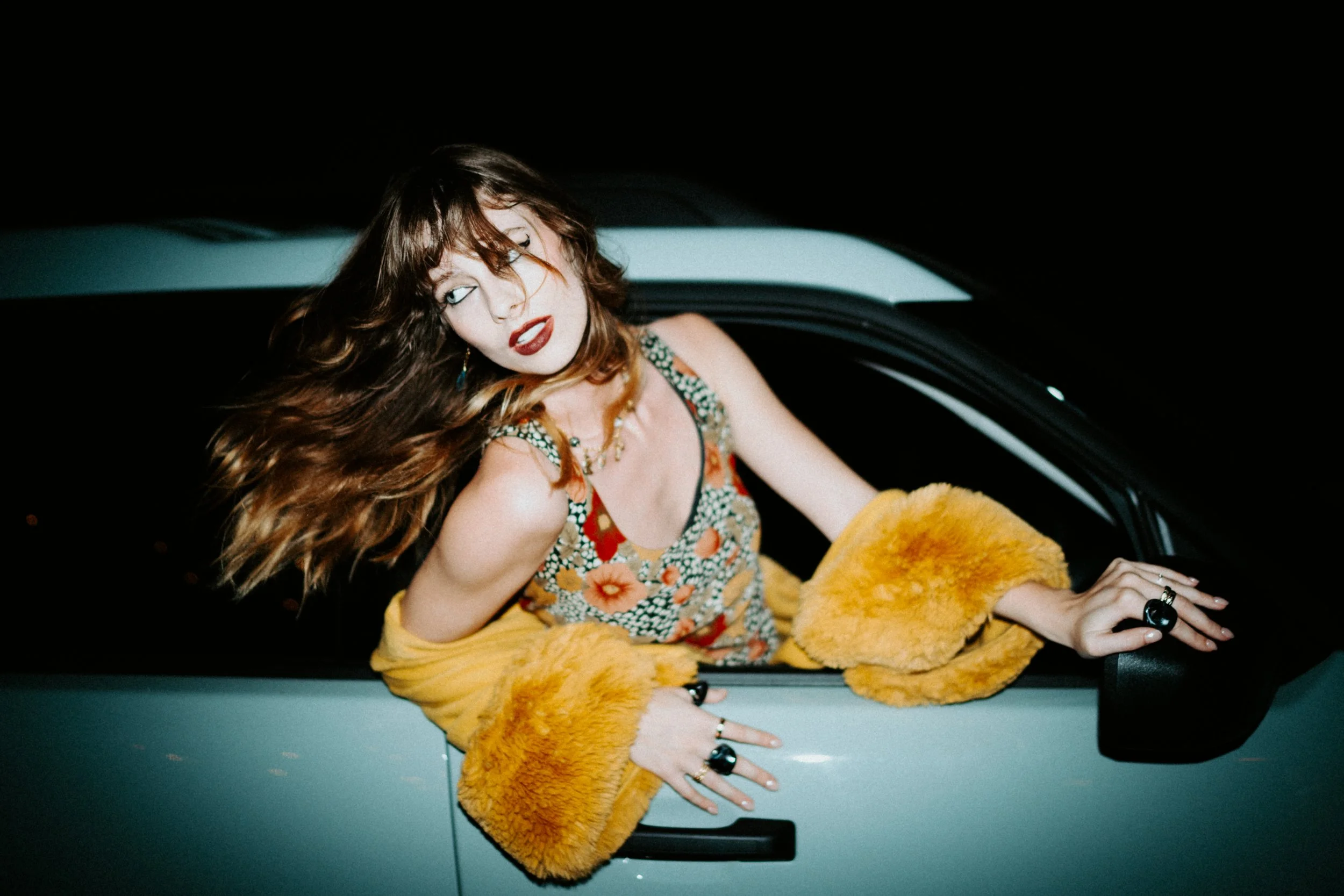 Woman with wavy hair and colorful dress leaning out of a car window at night, wearing rings and jewelry, with orange fuzzy coat draped over her arm.