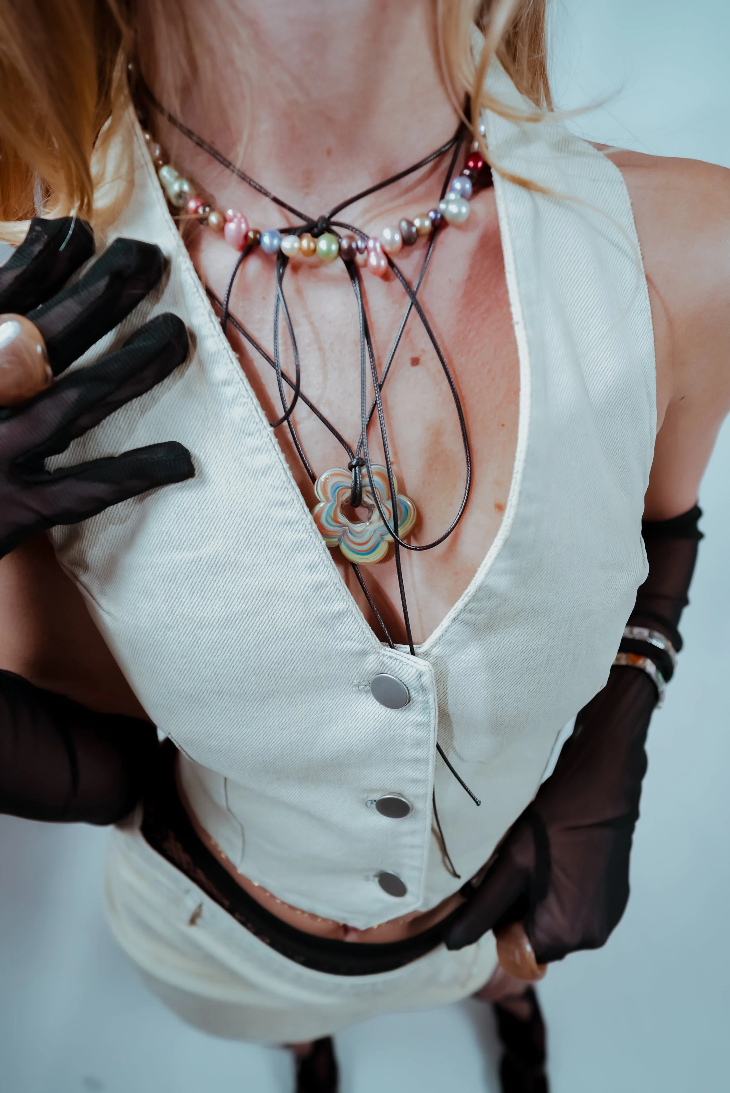 Close-up of a woman wearing multiple layered necklaces with colorful beads and a large flower-shaped pendant, a sleeveless white top with black ribbon accents, and black sheer gloves with rings. She has blonde hair and is photographed from a slightly above angle.