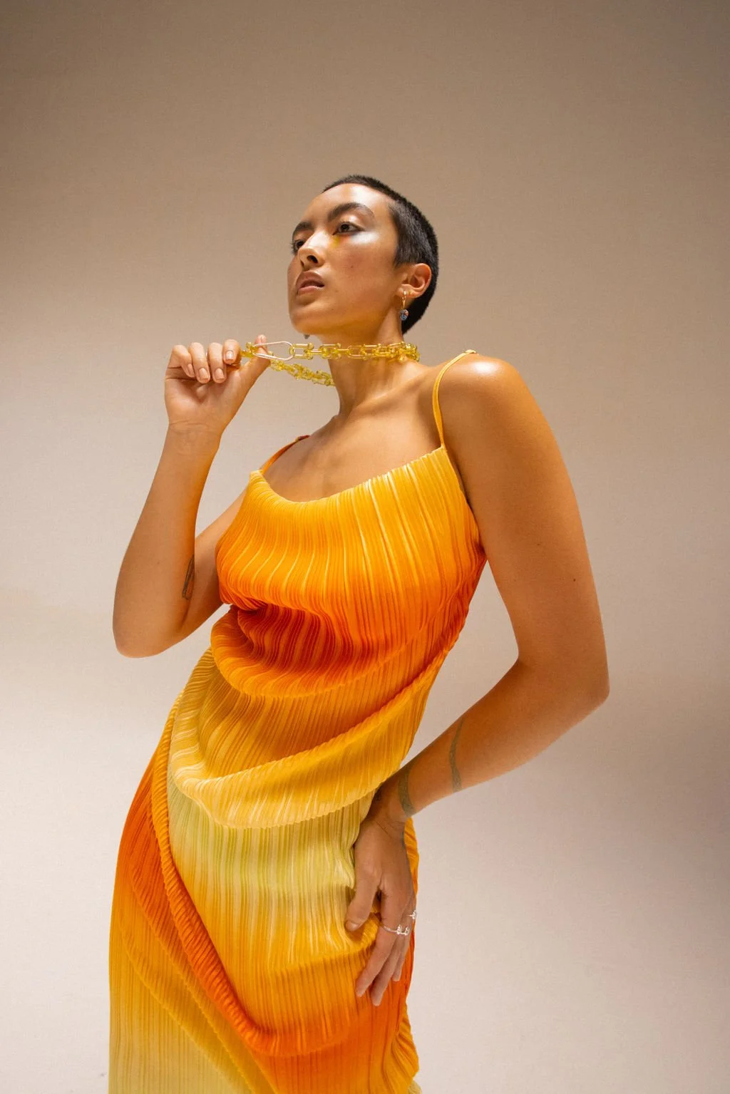 A woman with short dark hair and gold jewelry, wearing a vibrant orange and yellow pleated dress, posed against a plain background.