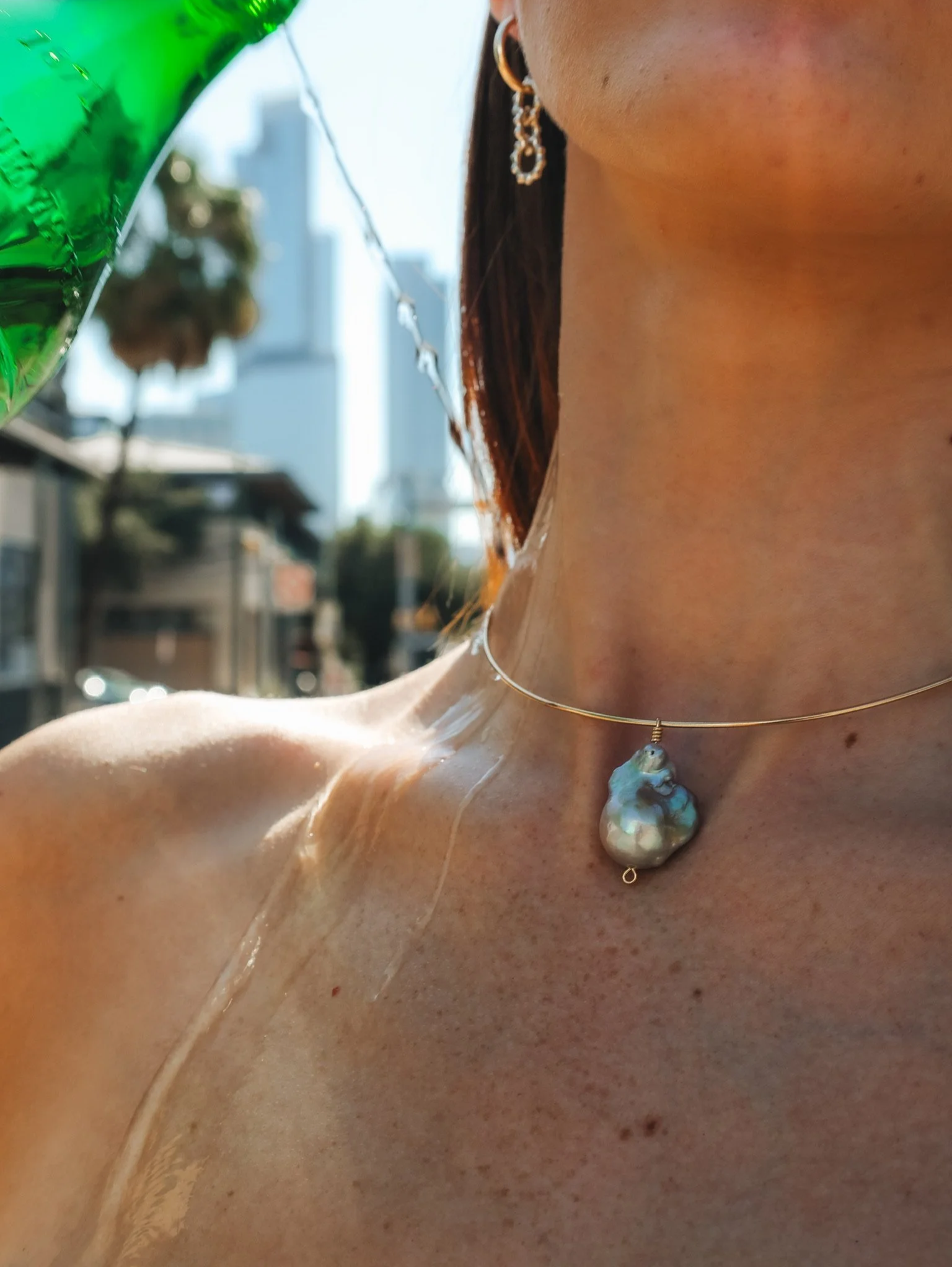 Close-up of a woman's neck and shoulder outdoors, wearing a pearl and shell necklace and a gold earring, with green balloons visible in the background.