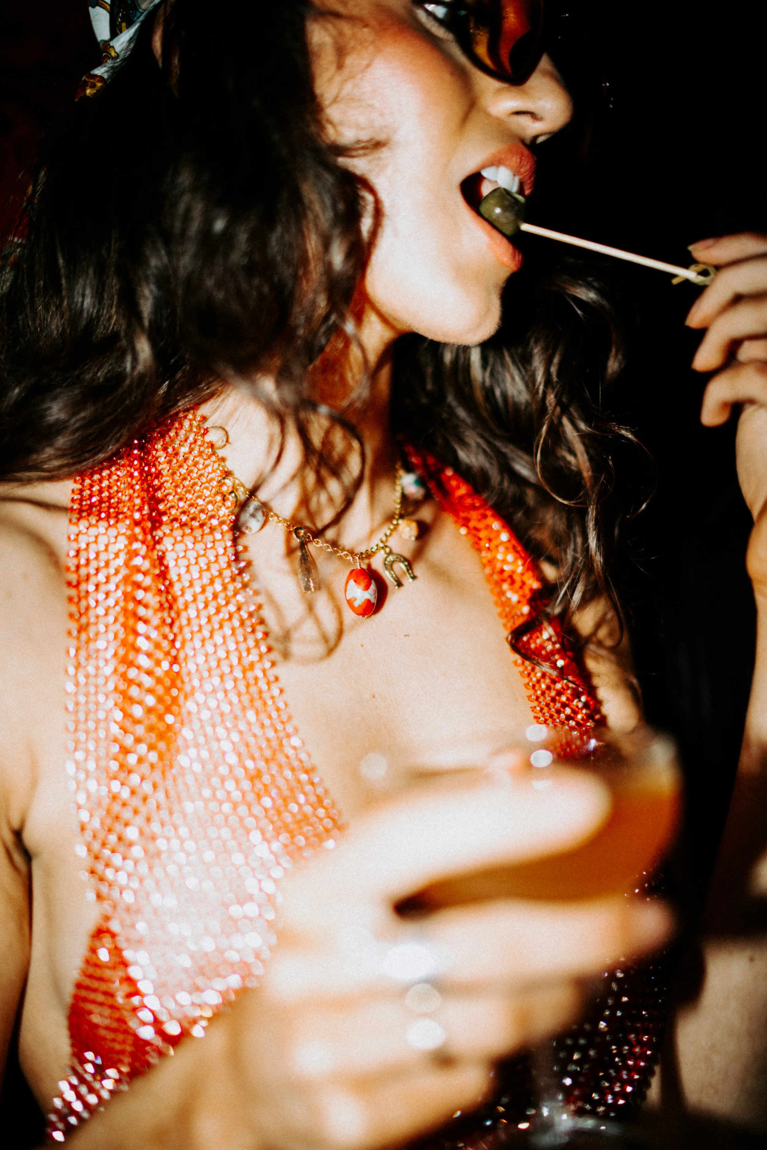A woman with dark, curly hair and orange lipstick, wearing sunglasses and a red beaded top, is holding a skewer with a green olive near her mouth, about to take a bite. She is also wearing necklaces.