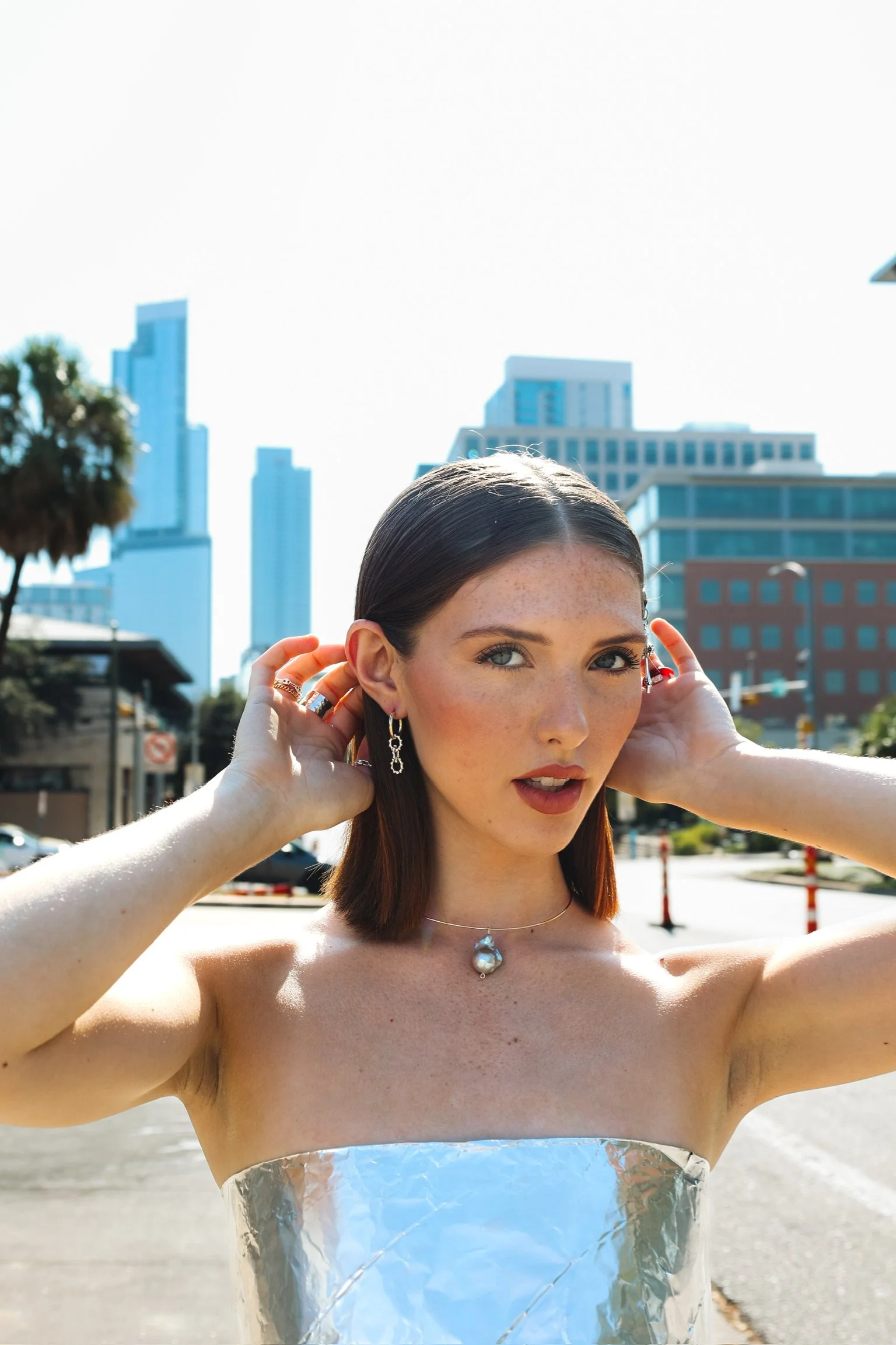 Young woman with dark hair posing outdoors in a cityscape background, wearing jewelry and a strapless top made of reflective material.