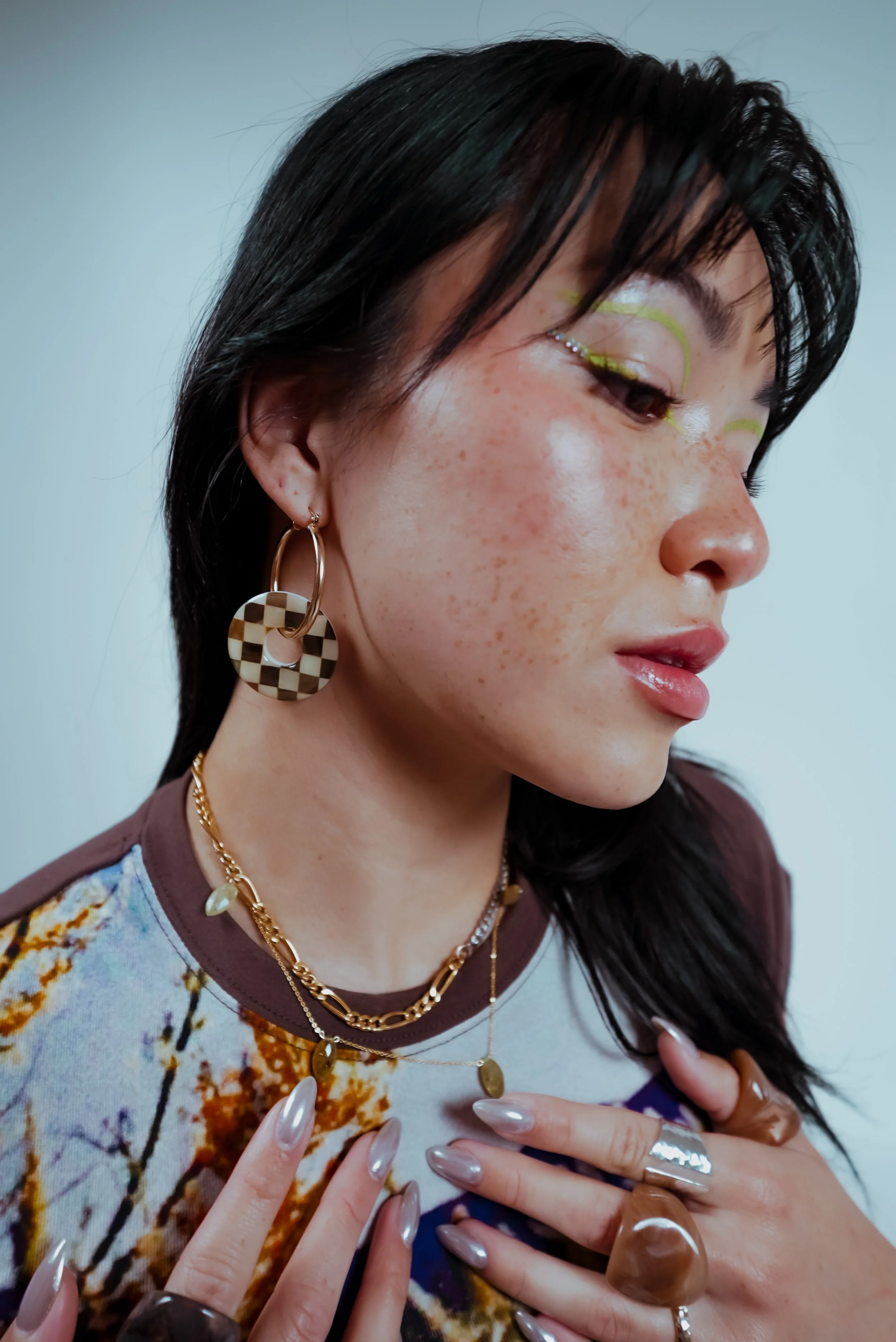 Close-up of a woman with black hair, wearing colorful makeup, gold earrings, layered gold necklaces, and multiple rings, with her hands near her chest and fingers extended.