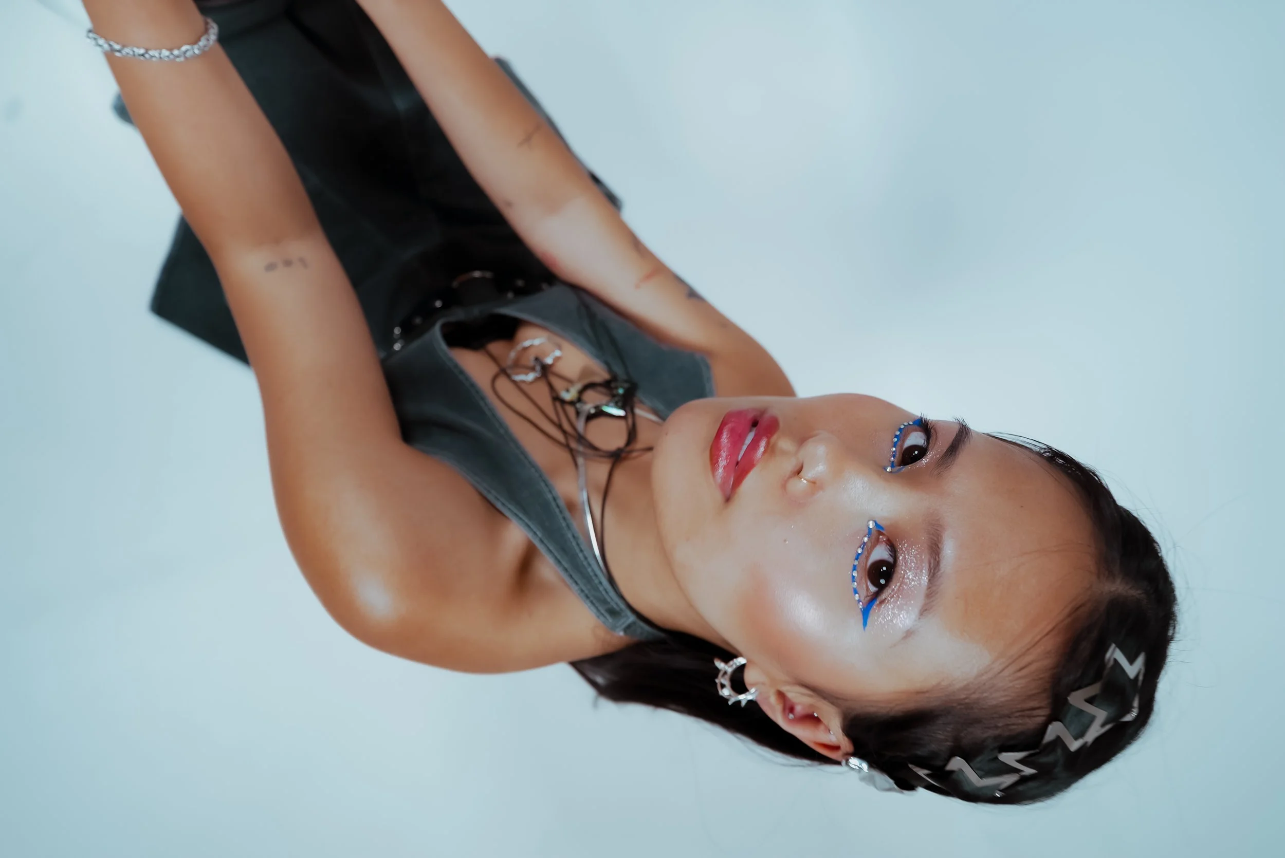 A young woman with short dark hair lying on her side against a plain light background, wearing a black sleeveless top with silver necklaces and earrings, and blue glitter makeup near her eyes.