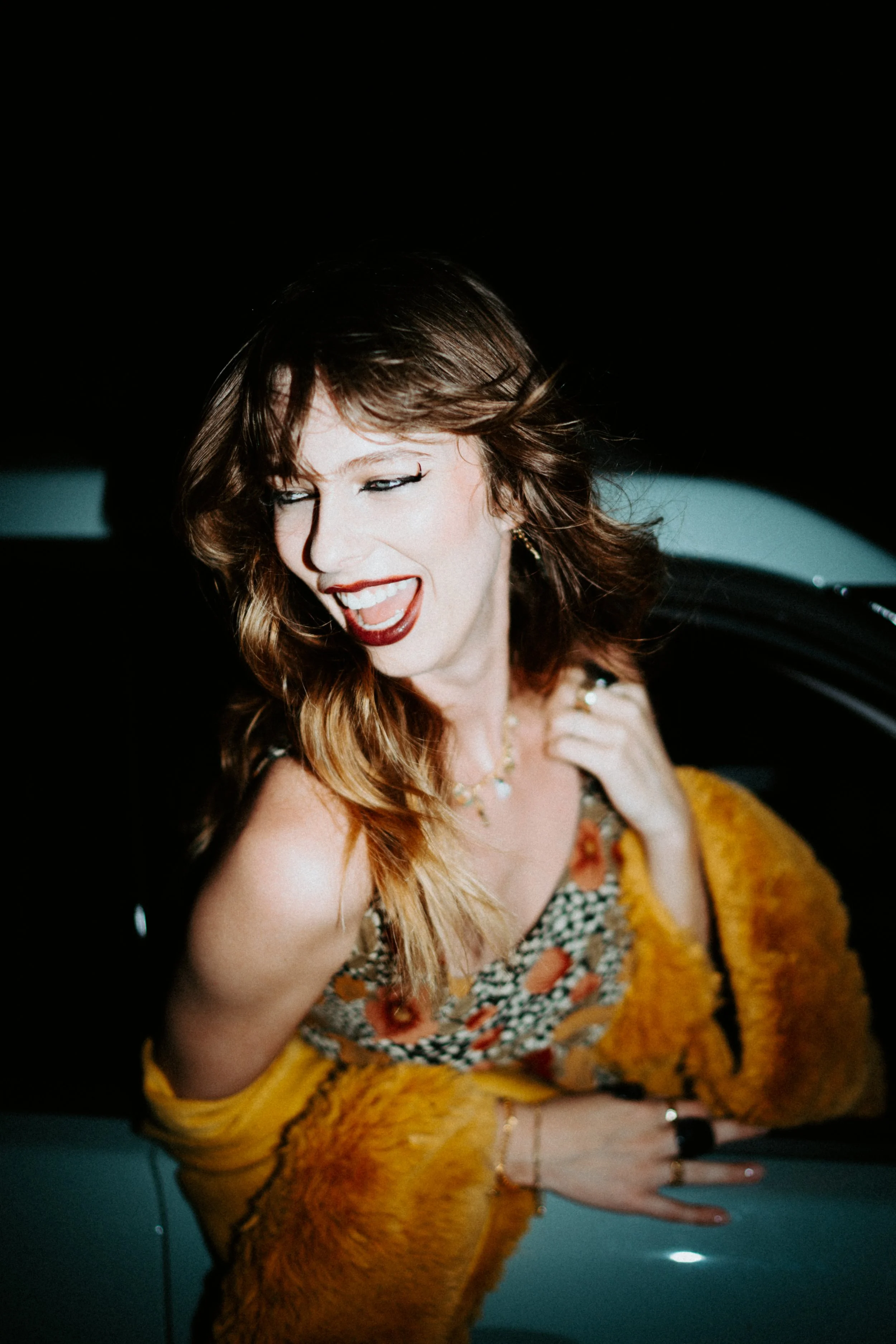 A woman with wavy brown hair singing with her eyes closed, wearing a colorful dress and yellow shawl, with jewelry, in front of a dark background.