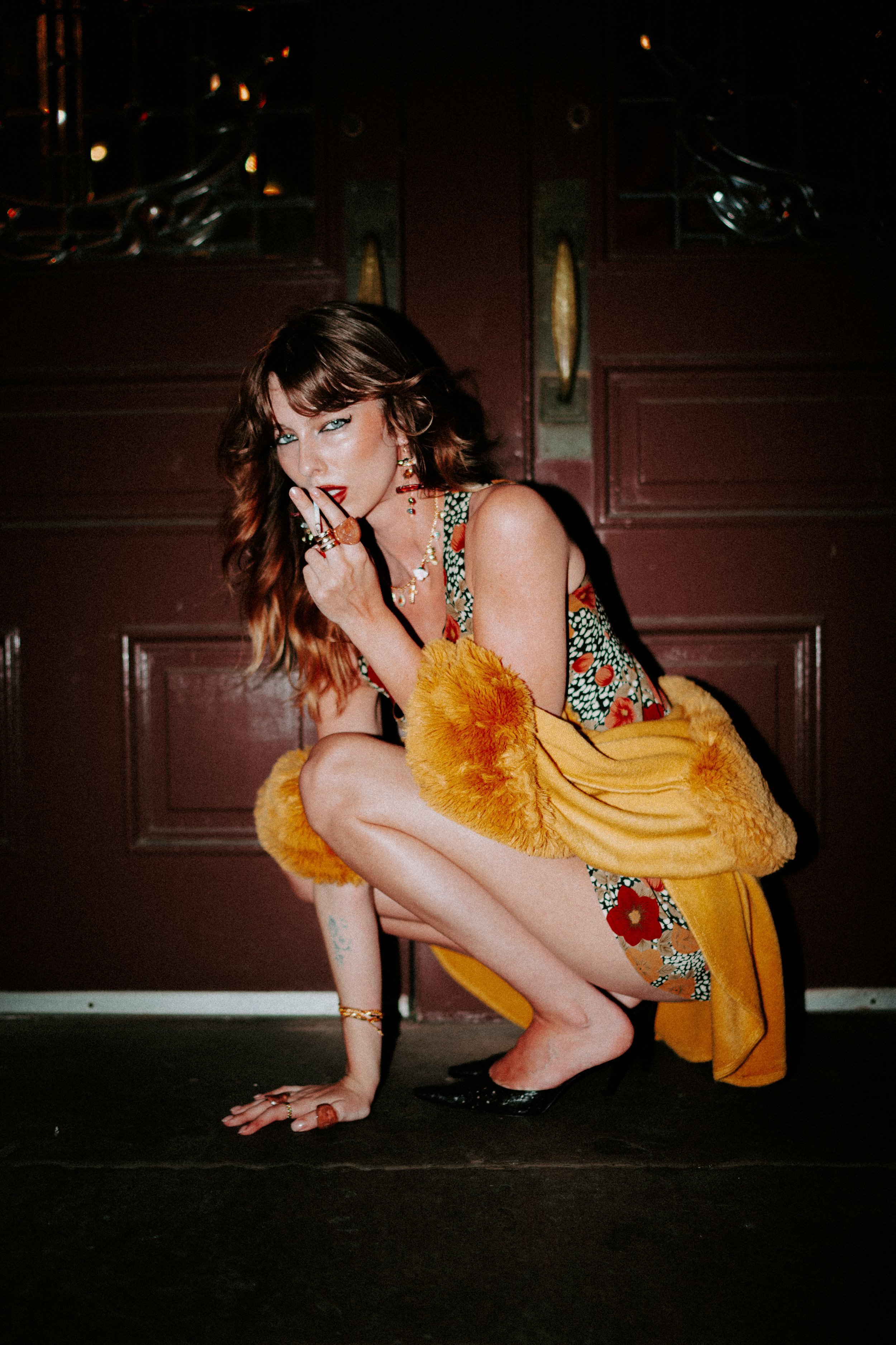 A woman with wavy brown hair and blue eyes, dressed in a colorful, patterned dress and a yellow fur coat, poses in a crouching position in front of a dark wooden door. She wears multiple rings, earrings, and necklaces, with bright red lipstick, and i