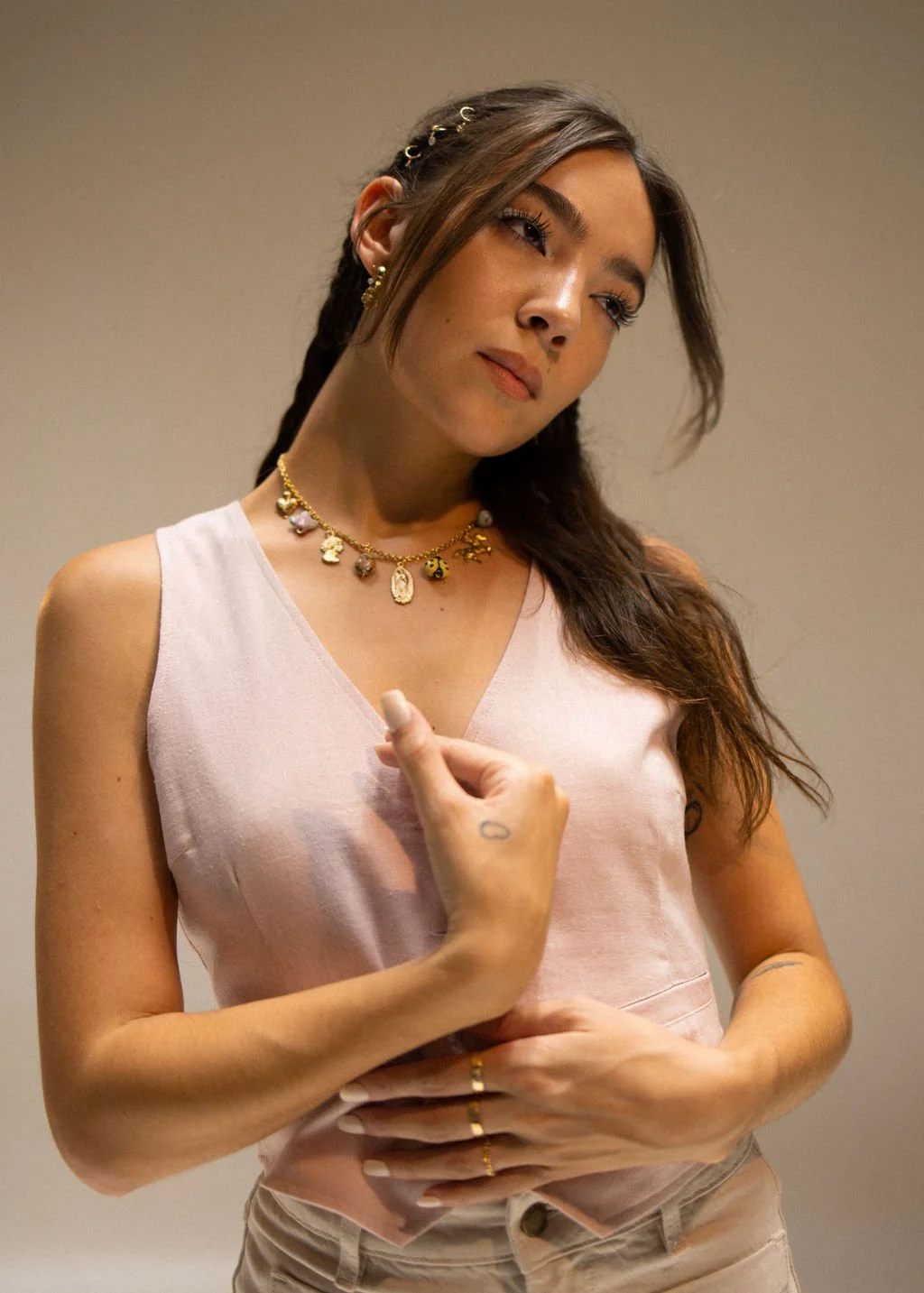 A young woman with long brown hair in braided style, wearing gold jewelry including necklaces and earrings, dressed in a light pink sleeveless top and beige pants, standing against a plain background.