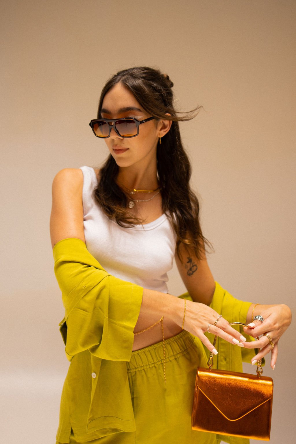 A woman with long brown hair wearing sunglasses, a white sleeveless top, and yellow cardigan and pants, holding a metallic orange purse, with jewelry, standing against a plain beige wall.