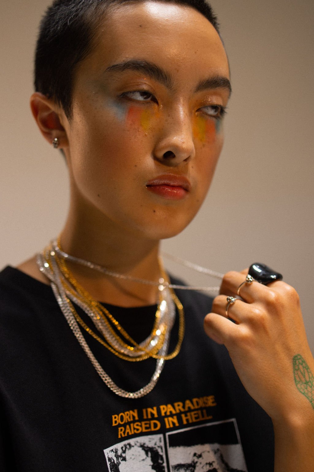 Close-up of a young person with short dark hair, colorful makeup, multiple gold and silver necklaces, rings, an earring, and a black T-shirt that reads 'BORN IN PARADISE RAISED IN HELL.' They are holding a chain with a large ring and have a green tattoo on their right hand.