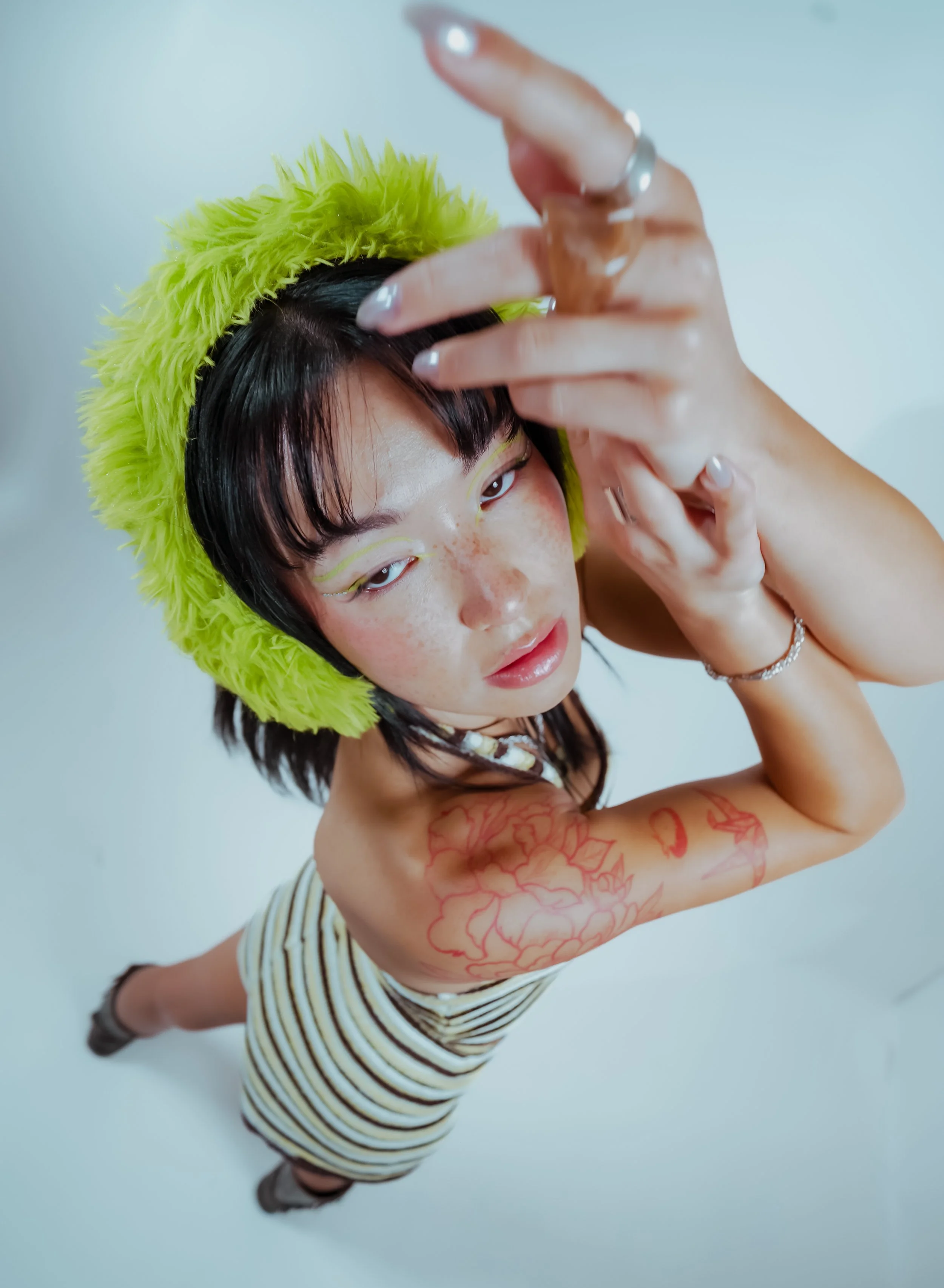 Person with short black hair wearing a neon yellow furry hat, eye makeup, and striped shirt looking at camera, holding their hand near face, standing against a white background.
