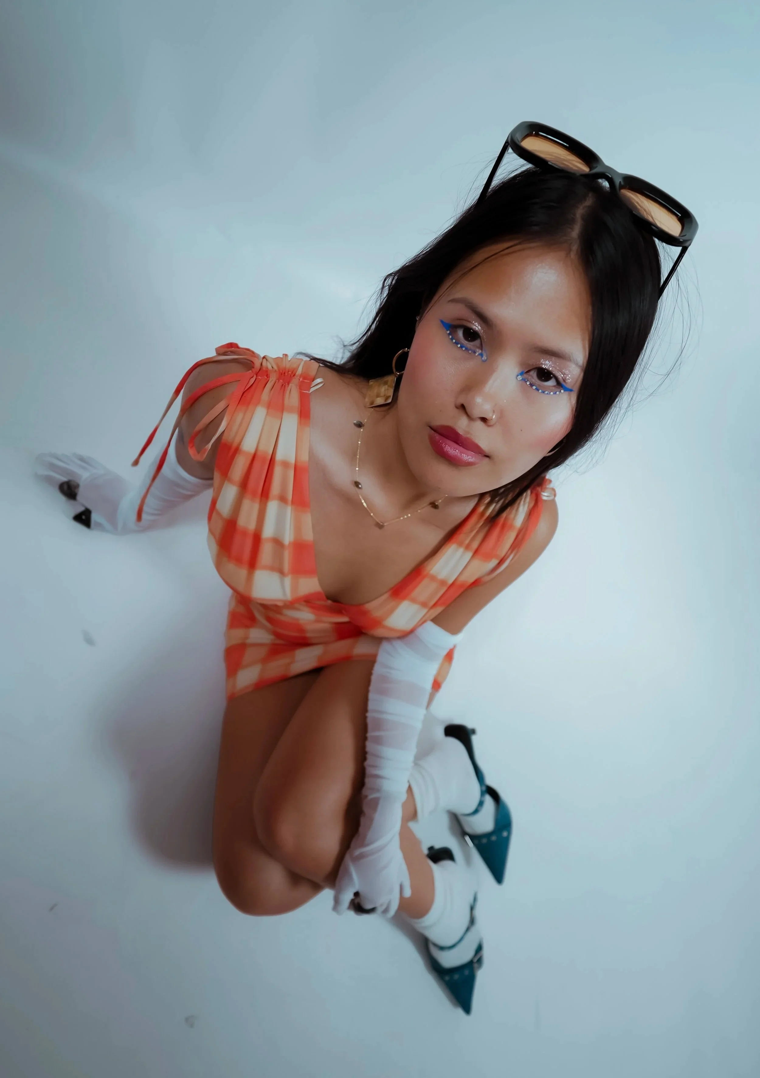 A woman with short black hair wearing orange checkered dress, white stockings, black heels, and glasses on her head, sitting on the ground with her legs crossed, looking at the camera in a studio with a plain white background.