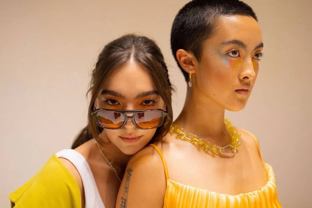 Two young women posing together against a neutral background, one with sunglasses and the other with bold makeup and jewelry.