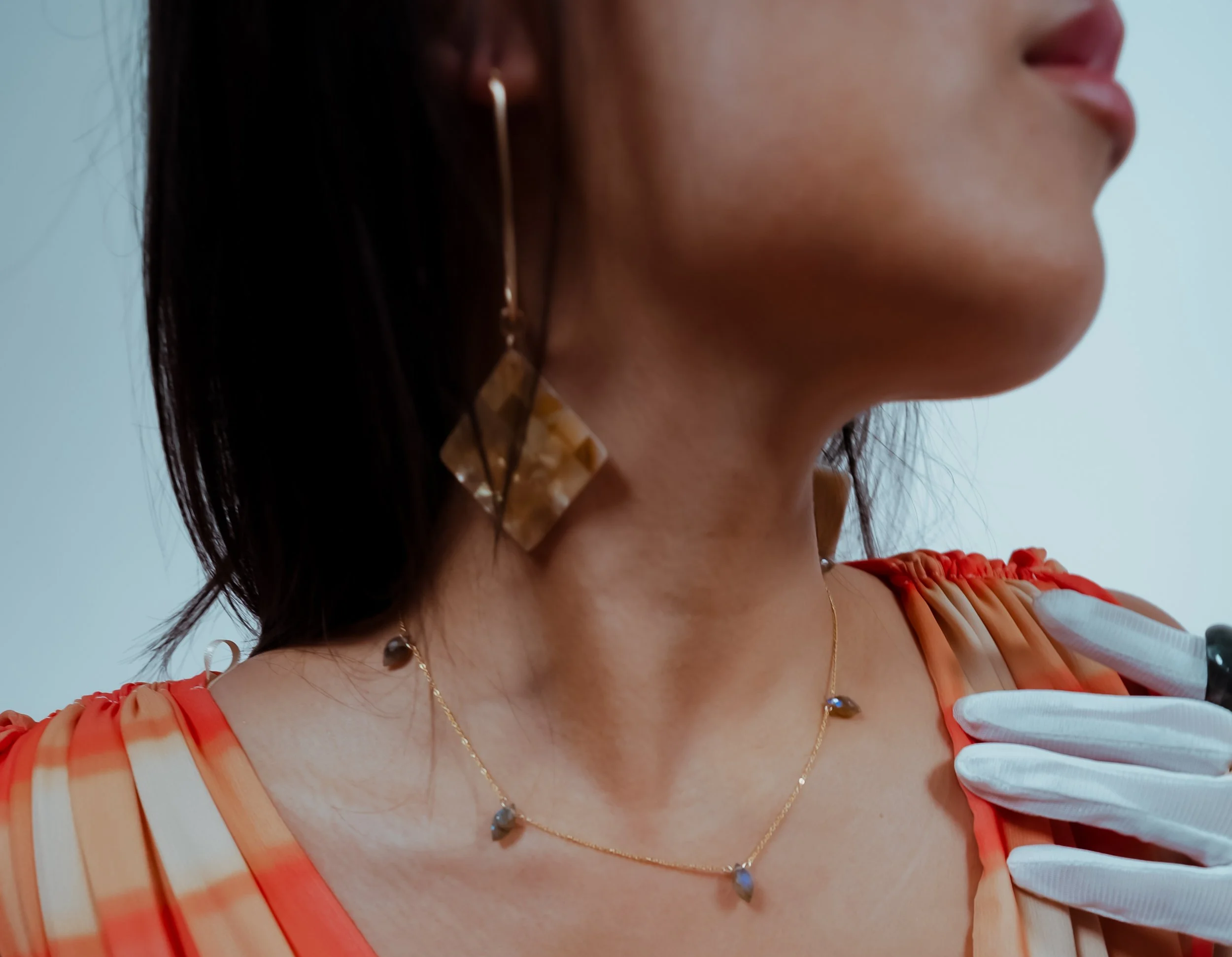 Close-up of a woman wearing a gold necklace with a dark stone pendant and a patterned strap top, smiling slightly, with dark hair, earrings, and a clear blue background.